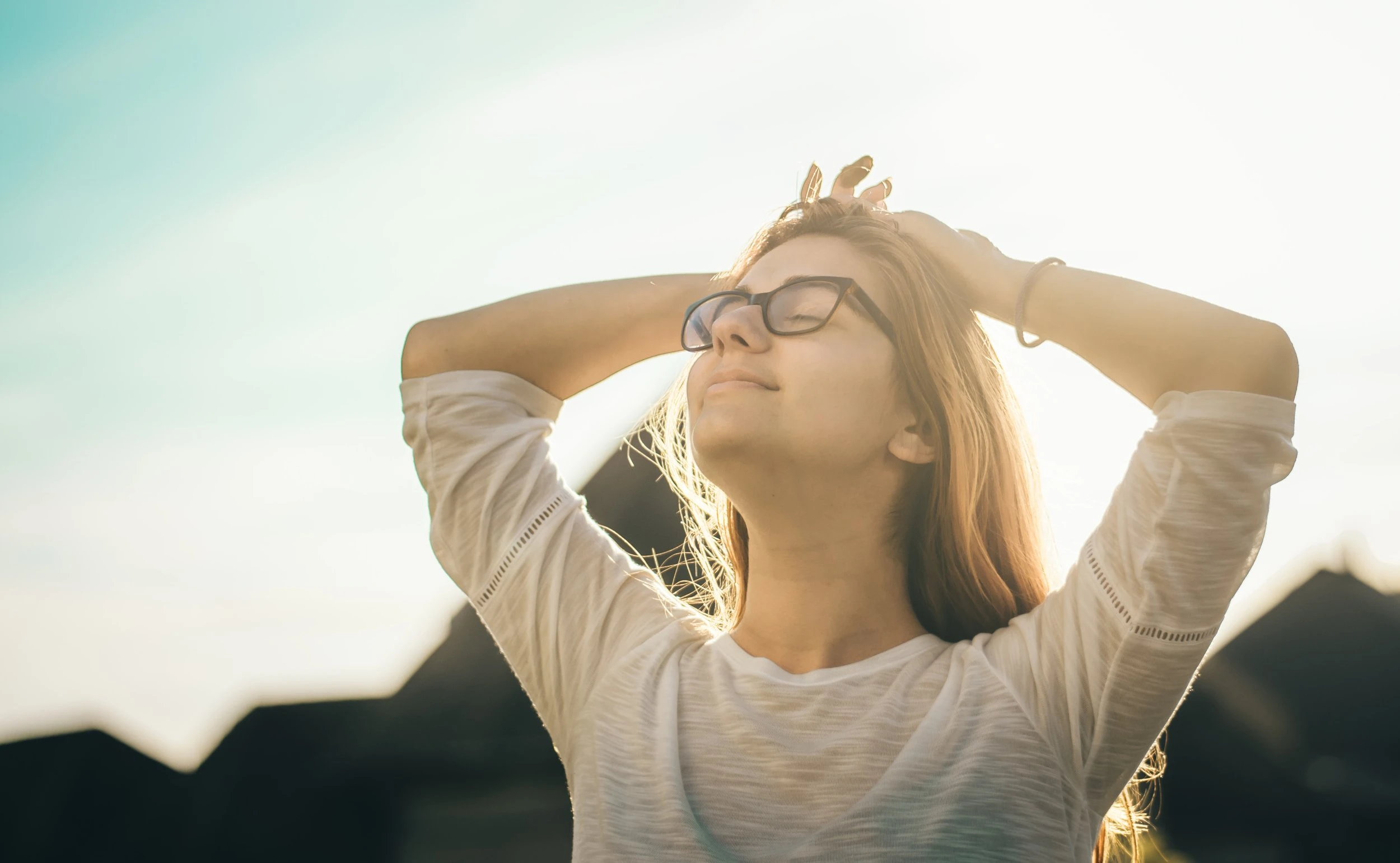 A young, blonde, female presenting person looking up to the sky with their eyes closed in relief and their hands resting on the top of their head. 