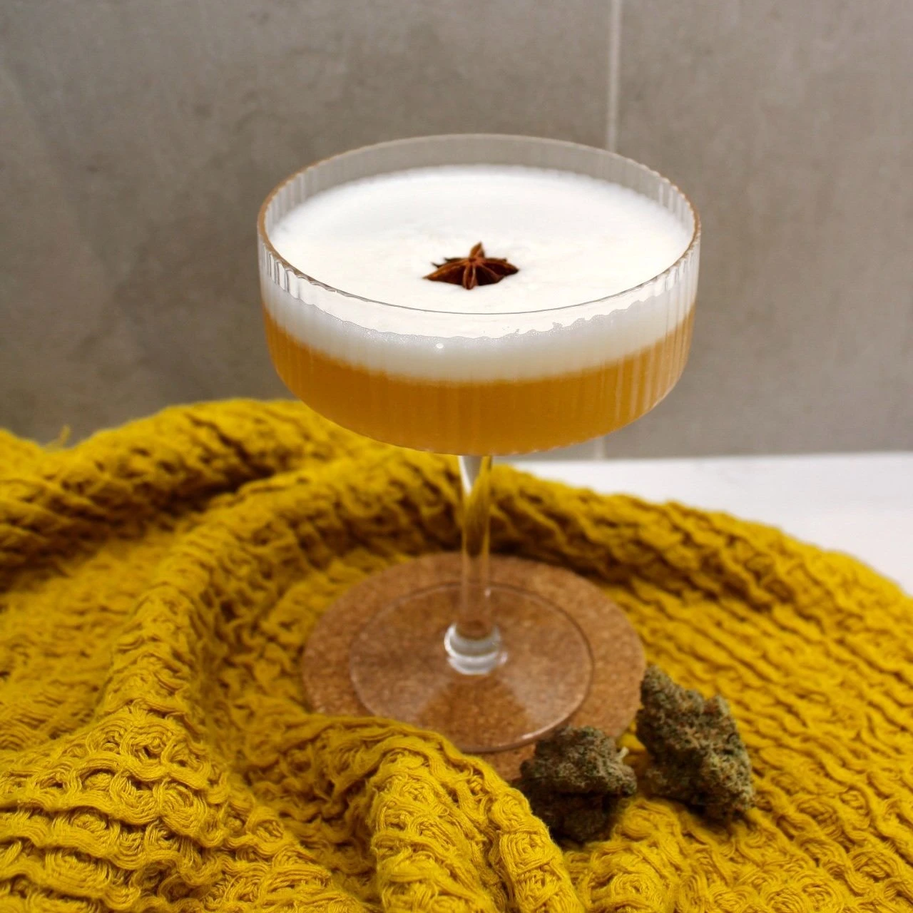 A martini glass full of a whiskey sour mocktail, garnished with a star anise and resting on a mustard yellow tea towel. In front of the glass sits two cannabis nuggets.