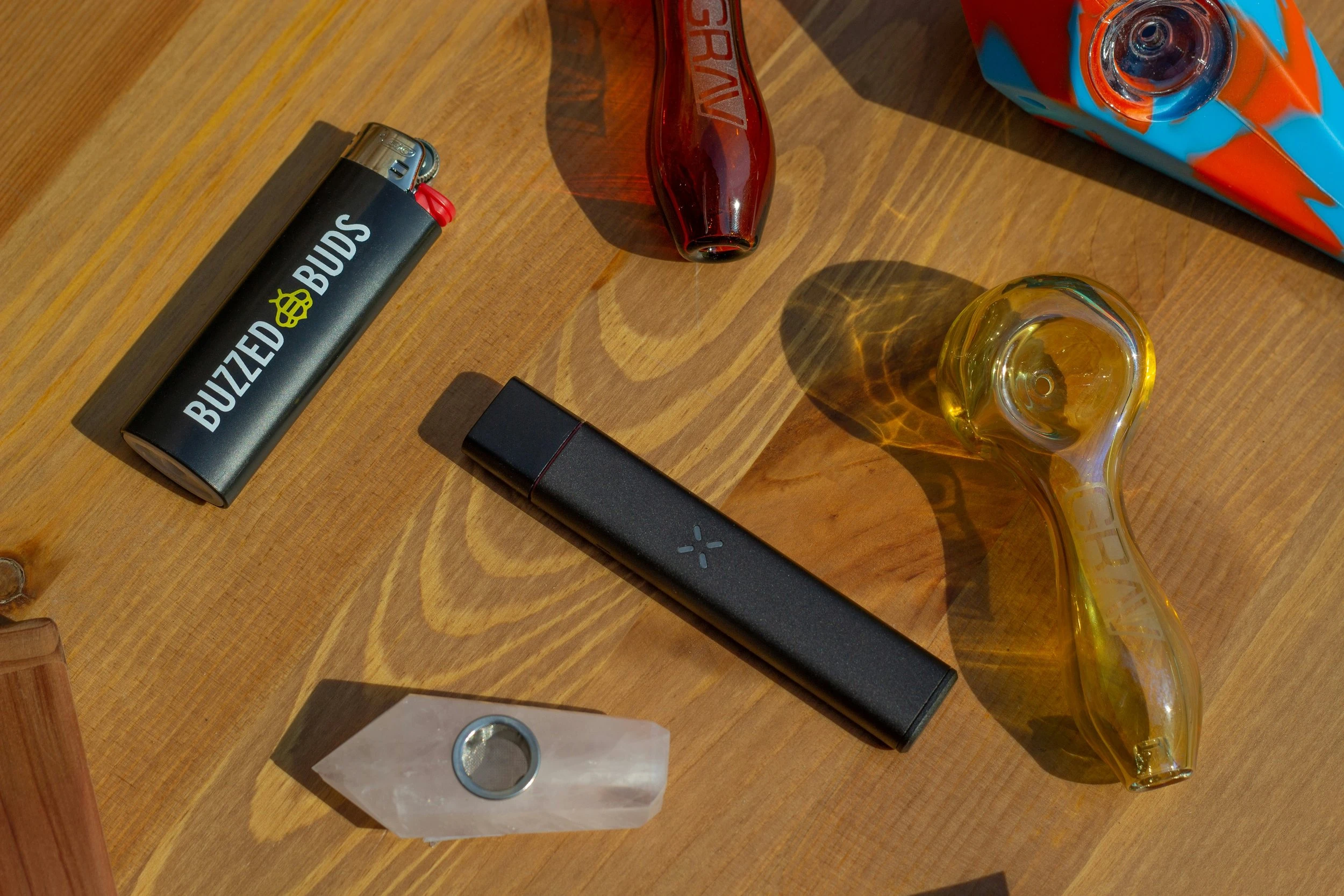 A bunch of cannabis tools includes pipes, vapes and lighters all lying on a light wooden surface and are skewed about.