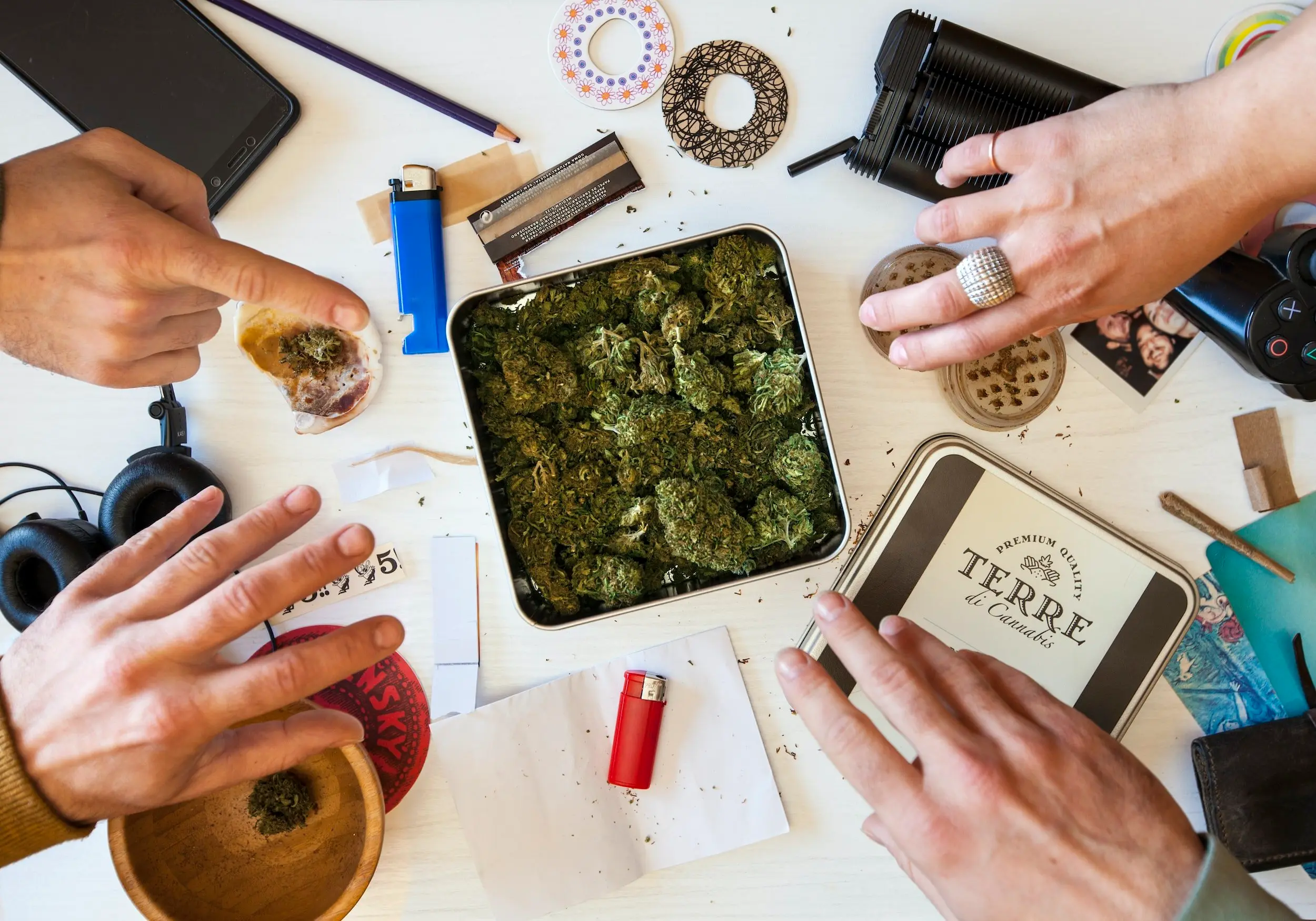 A large square tin of cannabis nuggets surrounded by other cannabis/stoner tools like lighters, grinders and rolling papers.