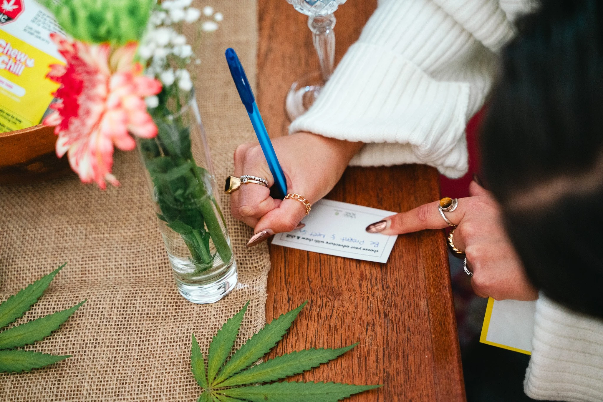 A card rests on a wooden table with flowers and cannabis leaves surrounding t, as a woman with long gold nails writes something down on it with a blue pen.