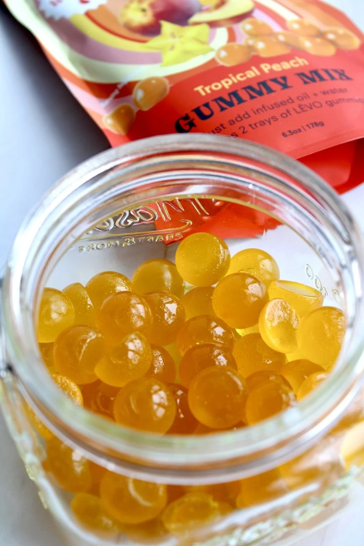 A clear mason jar full of small peach coloured, round gummies. Behind the jar is a package of tropical peach gummy mix.