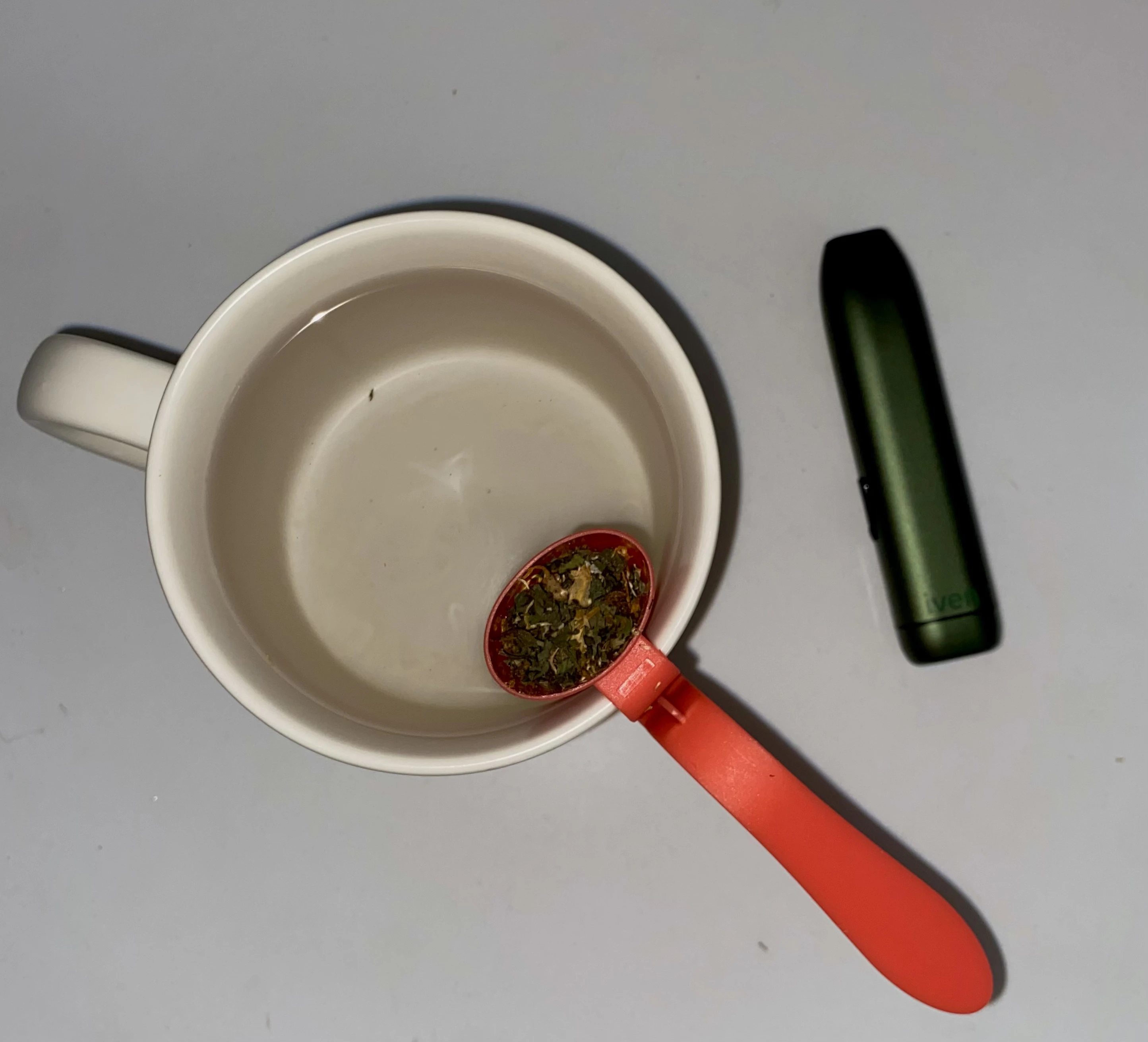 An aerial view of a beige mug full of hot water and a pink tea steeper on the side, infusing resting beside the camp green Iven dry herb vaporizer.