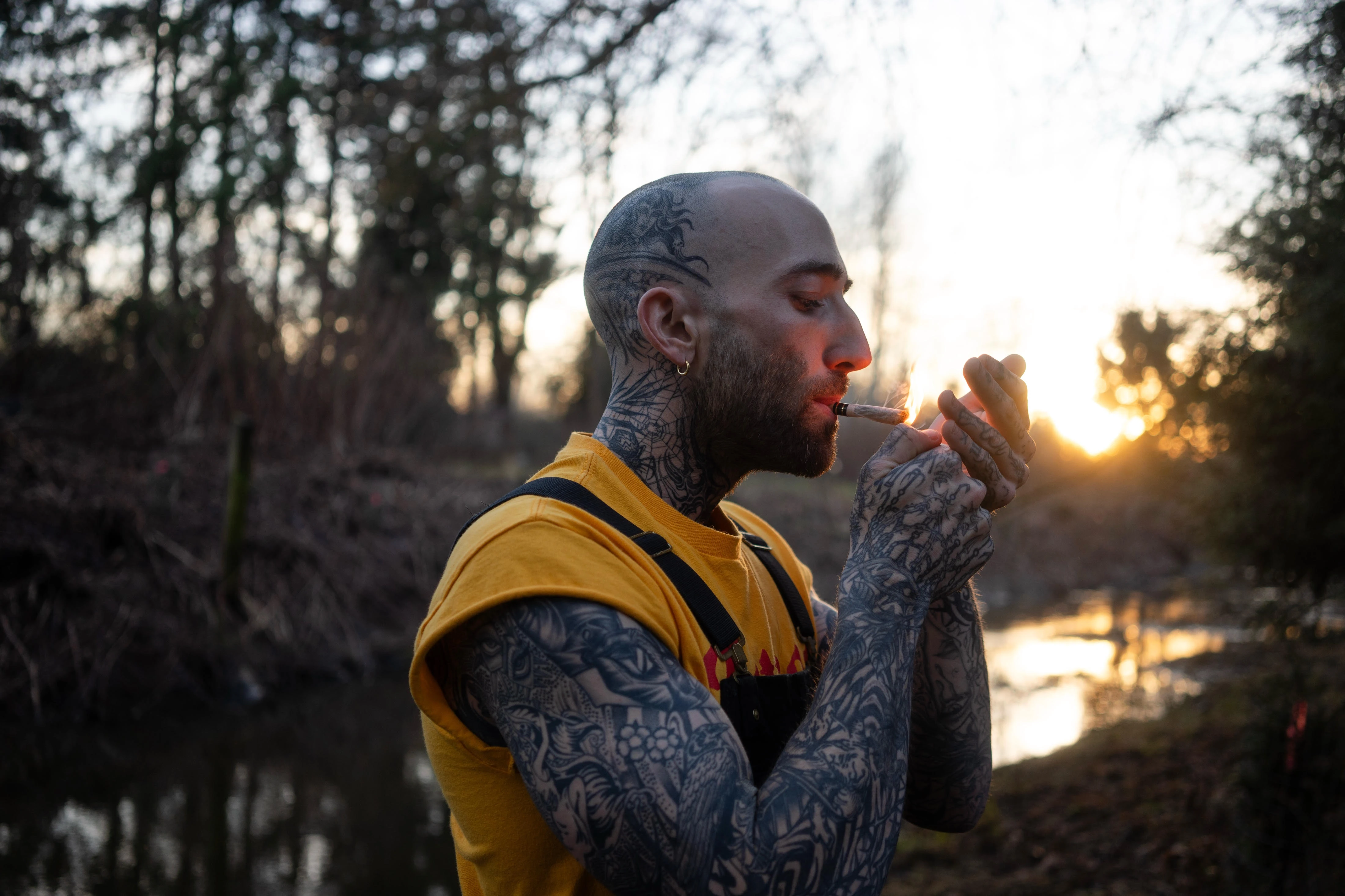 tattooed man in a yellow t-shirt smoking a joint in the forest at sunset