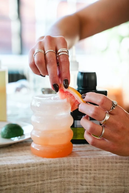 A bubble glass full of an ombre pink grapefruit mocktail, and two hands are adding a grapefruit wedge on to the side of the glass as a garnish. 