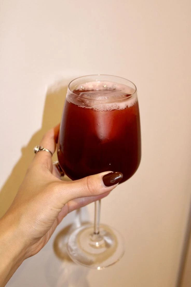 A wine glass holding a cranberry based mocktail inside of it, which is held up by a hand with red, long fingernails and a silver ring.