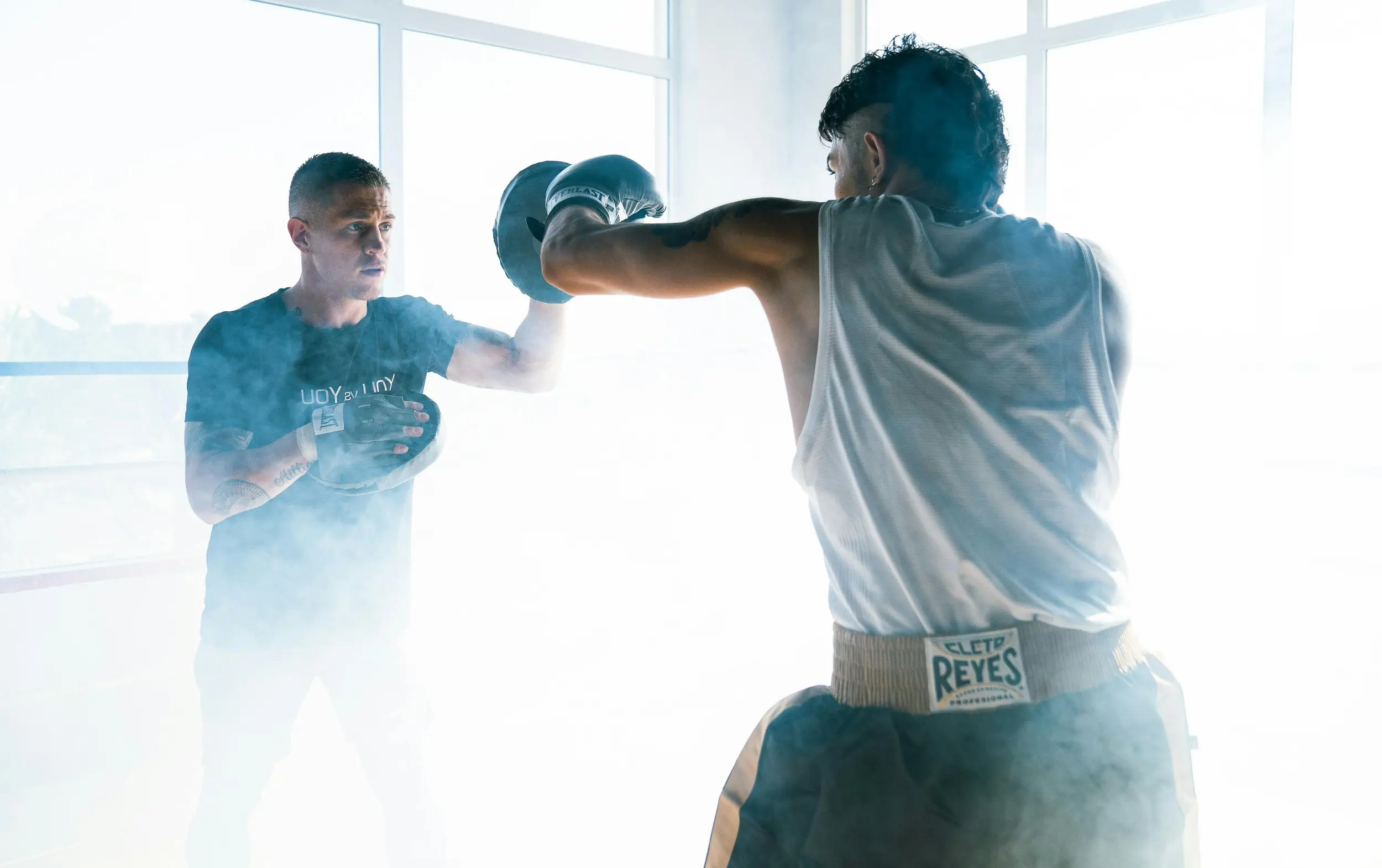 Two men practicing boxing. One of them is wearing gloves and the other is holding padded targets for the guy to hit. They are enveloped in clouds of smoke and dust.