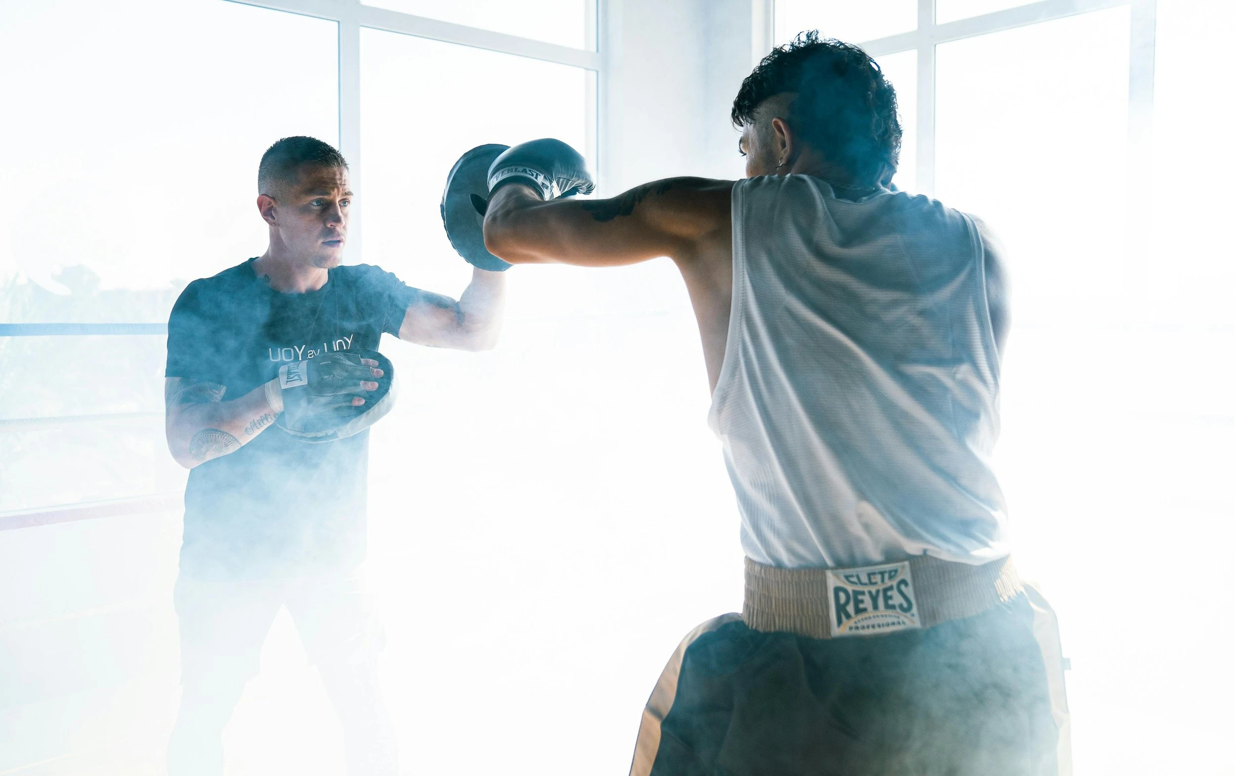 Two men practicing boxing. One of them is wearing gloves and the other is holding padded targets for the guy to hit. They are enveloped in clouds of smoke and dust.