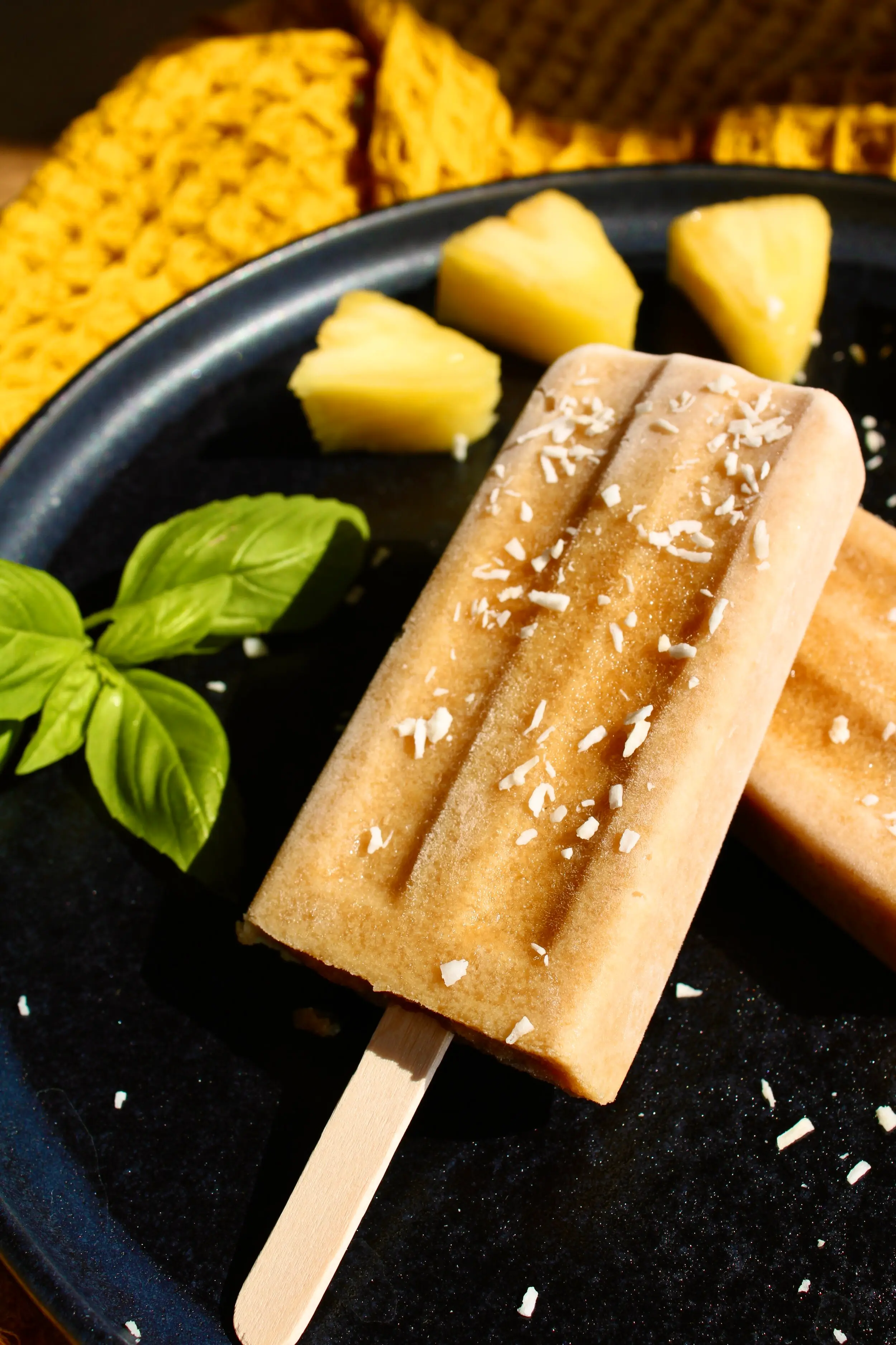 A brown/orange coloured popsicle on a black plate surrounded by sprigs of mint and chunks of pineapple.