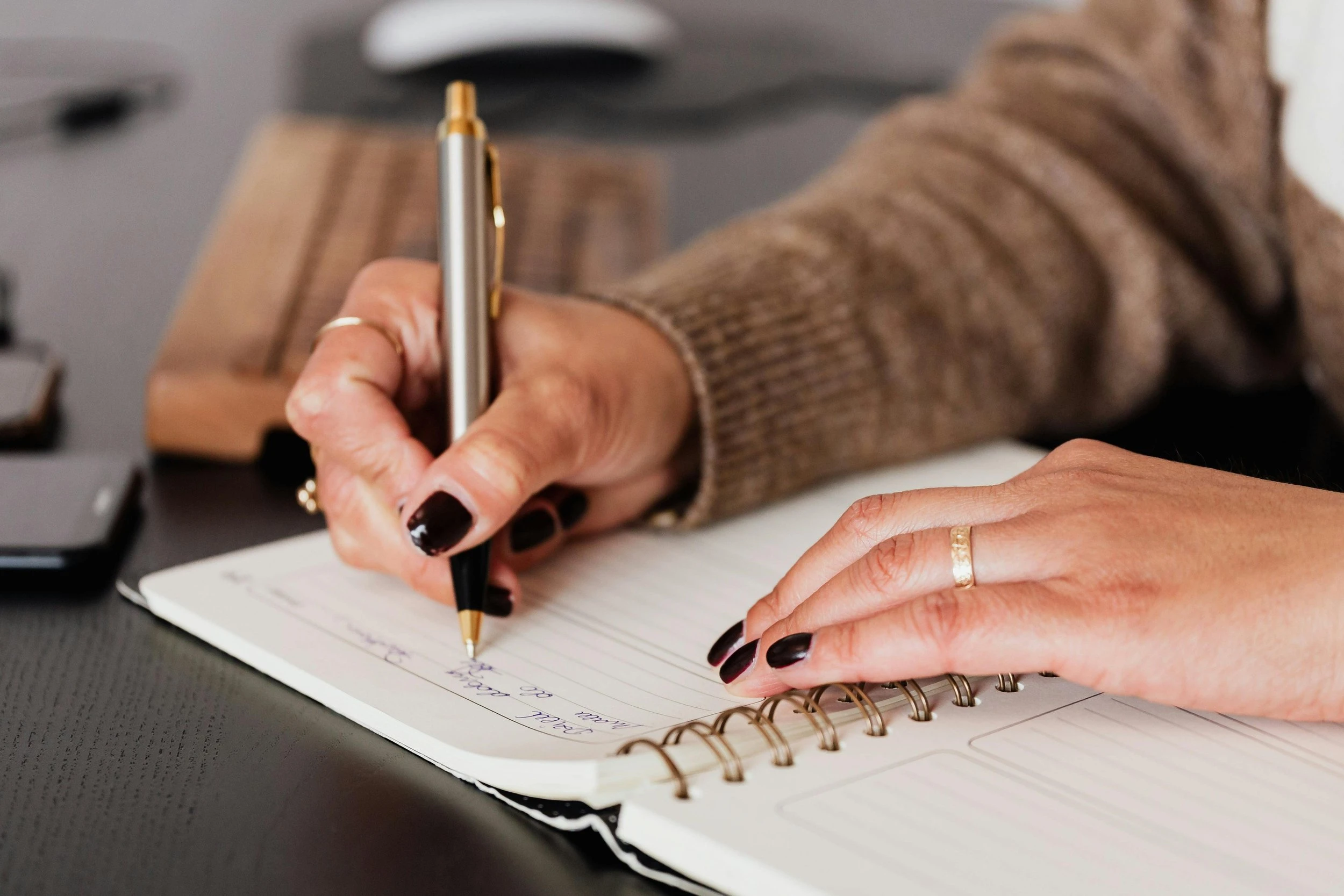 A hand with black nail polish holding a gold pen and scribbling on a notepad.