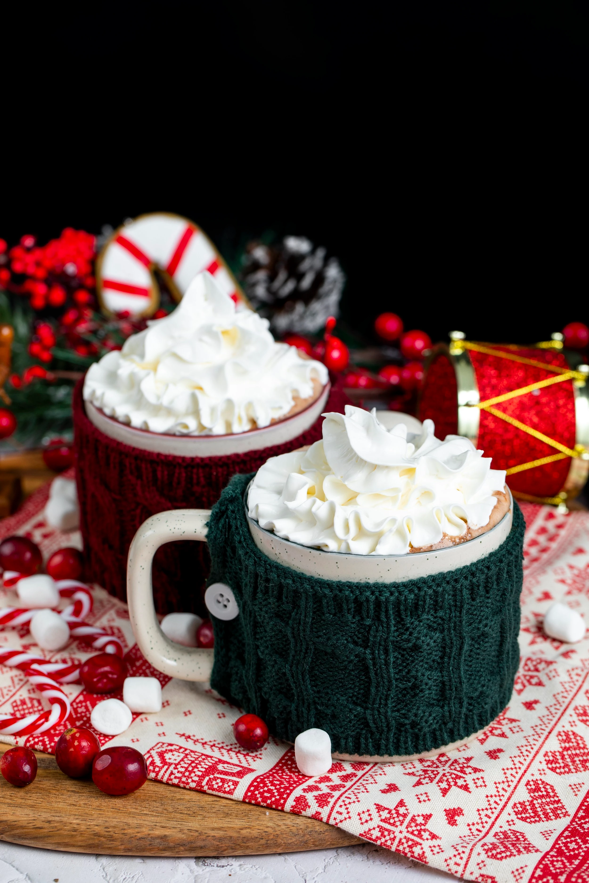 Two mugs of hot chocolate, overflowing with whipped cream. One mug has a green knit cozy over it while the other has a red knit cozy. 