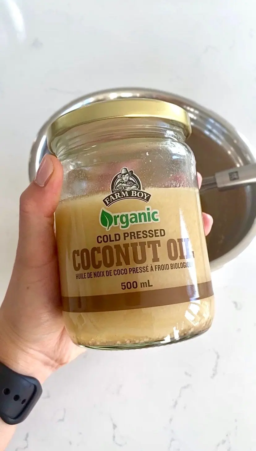 a hand holding up a large, clear jar of farm boy coconut oil.