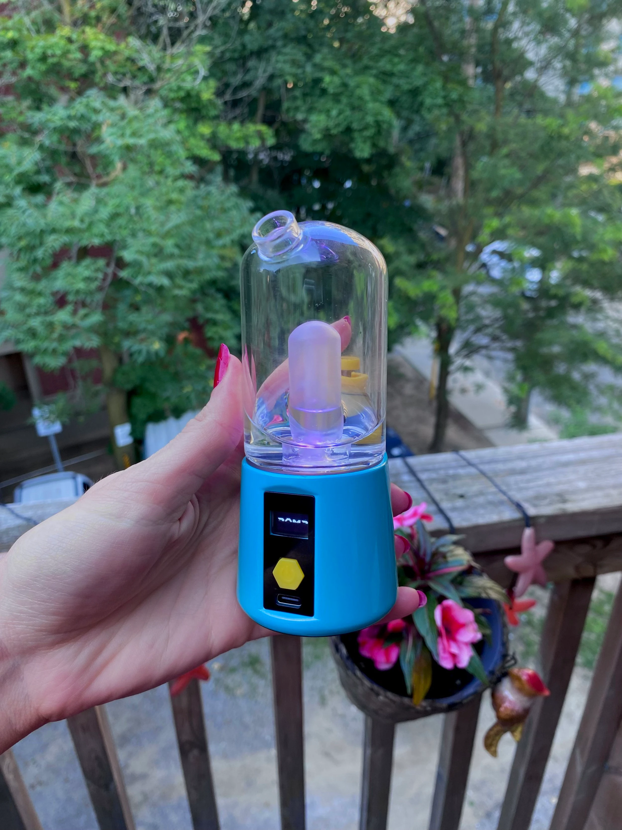 A hand with red nails holding the teal coloured Bomb electric dab rig on a balcony.