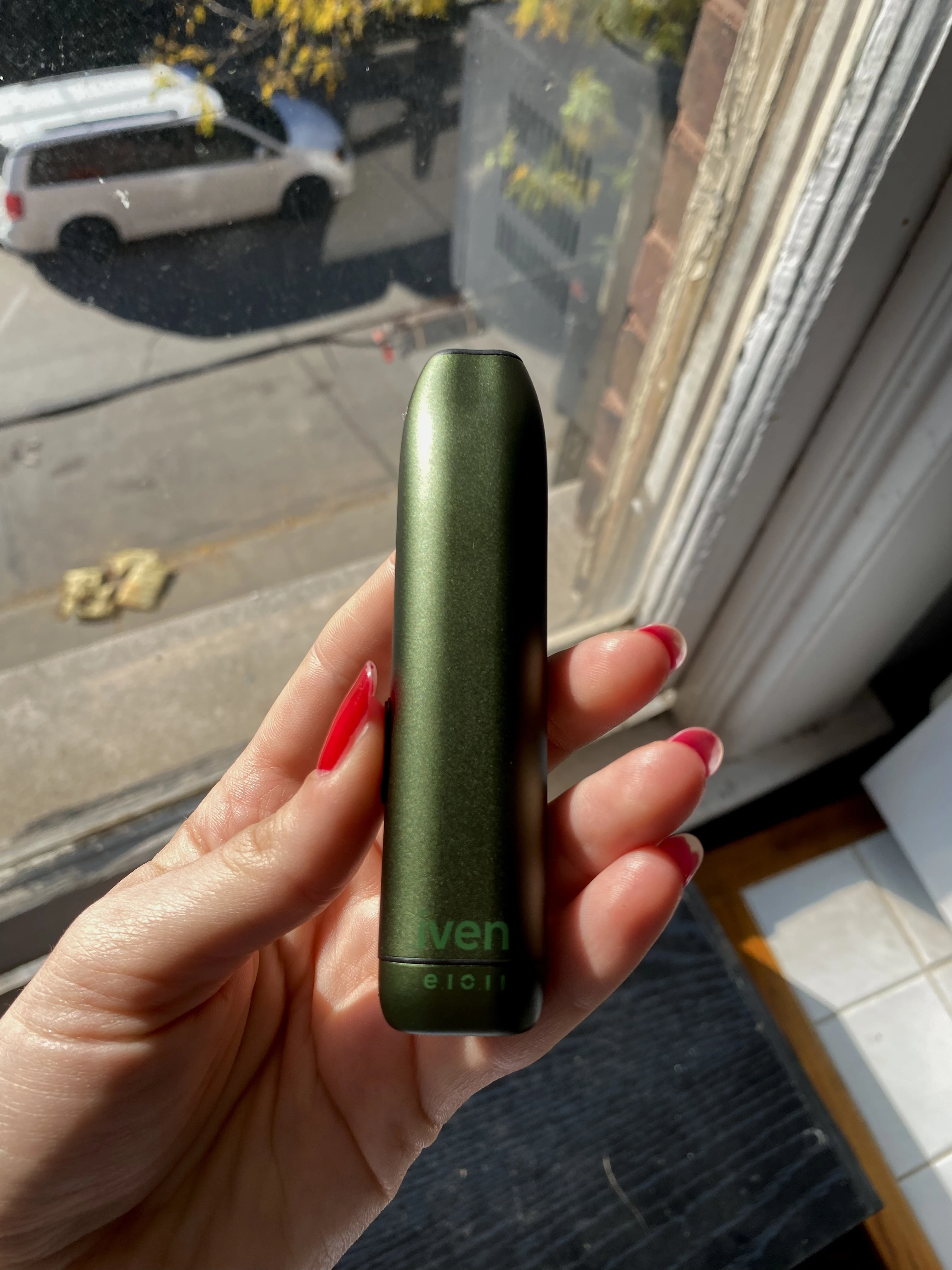 A hand with red finger nails holding up the olive green Iven dry herb vaporizer in the sunlight next to a large window. 