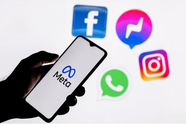 A hand holding a cellphone with the Meta logo on it with the Instagram, Facebook, WhatsApp and Messenger floating around the rest of the image. 