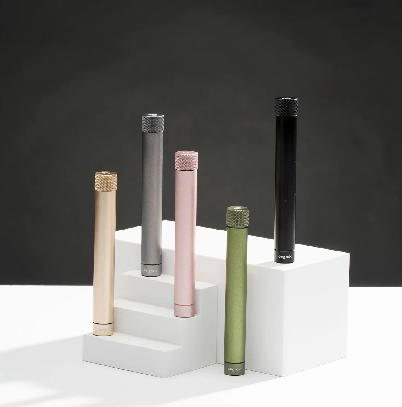 A pink, green, silver, black and gold Ongrok cannabis joint holder stand on white blocks against a charcoal black backdrop.