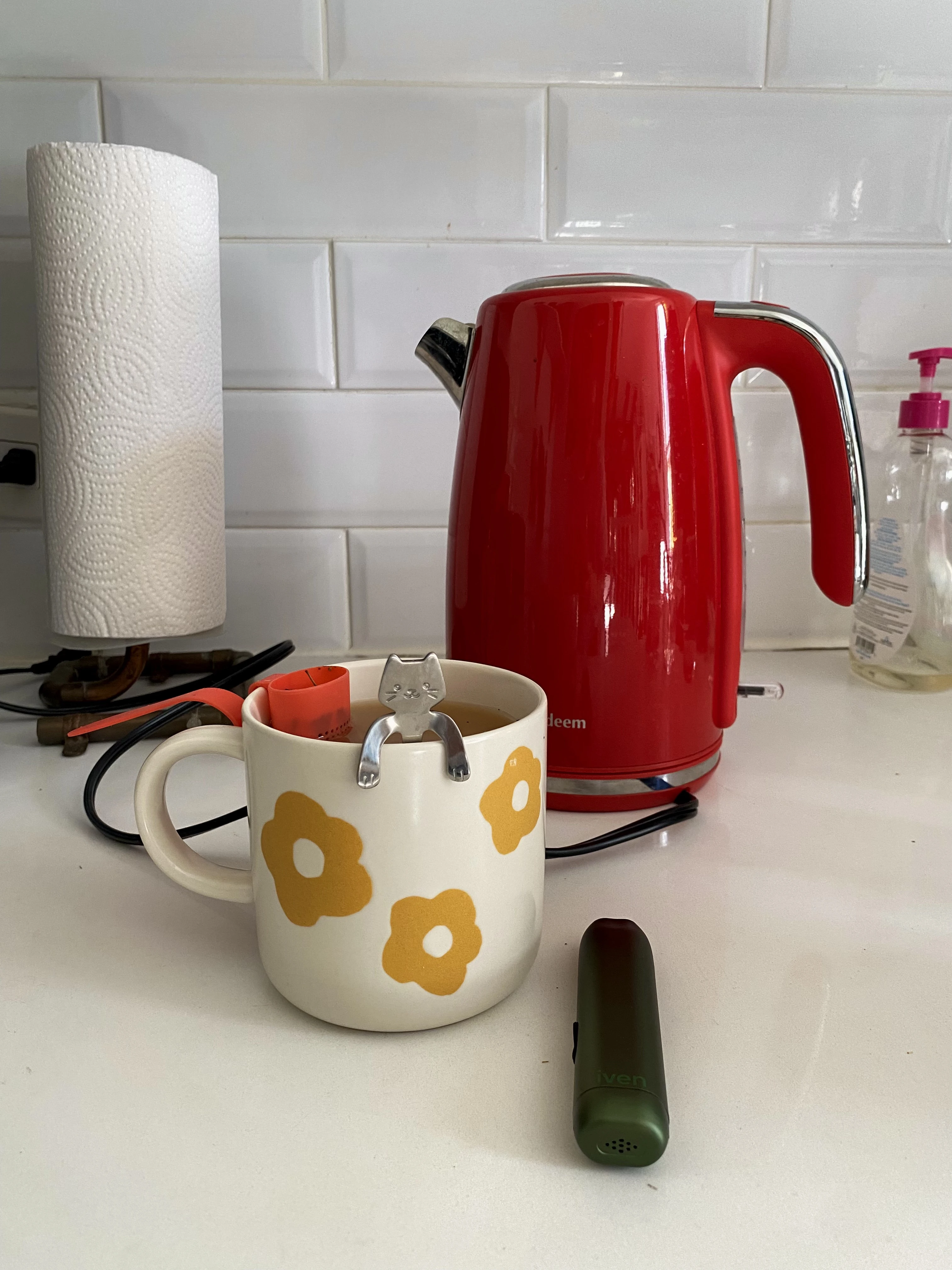 A beige mug with orange flowers on it rests next to a bright red kettle and the camp green Iven device as a pink steeper inside the mug steeps the tea. 