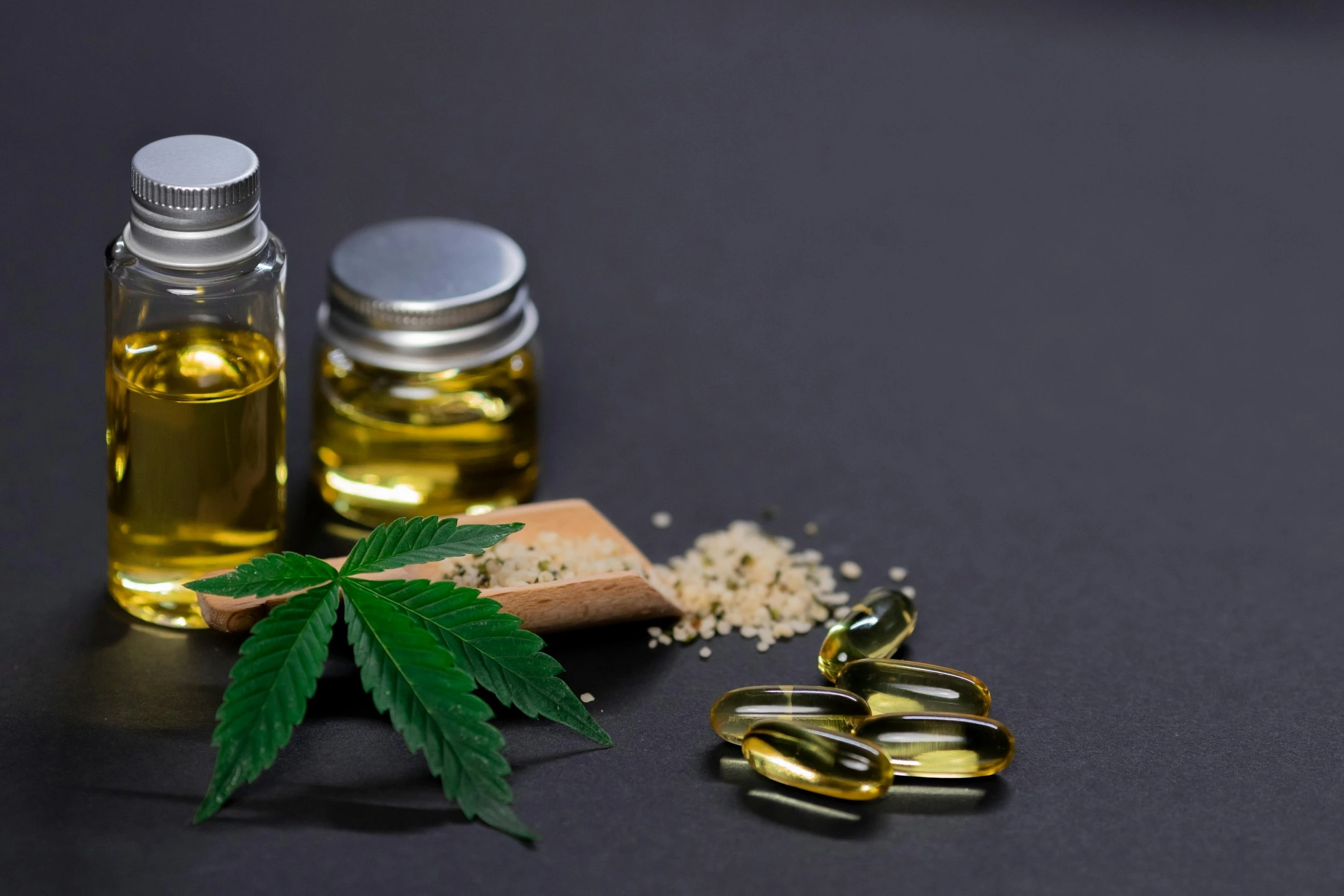 Two jars of cannabis oil, four gel capsules and a green cannabis leaf rest on a charcoal grey counter and backdrop.