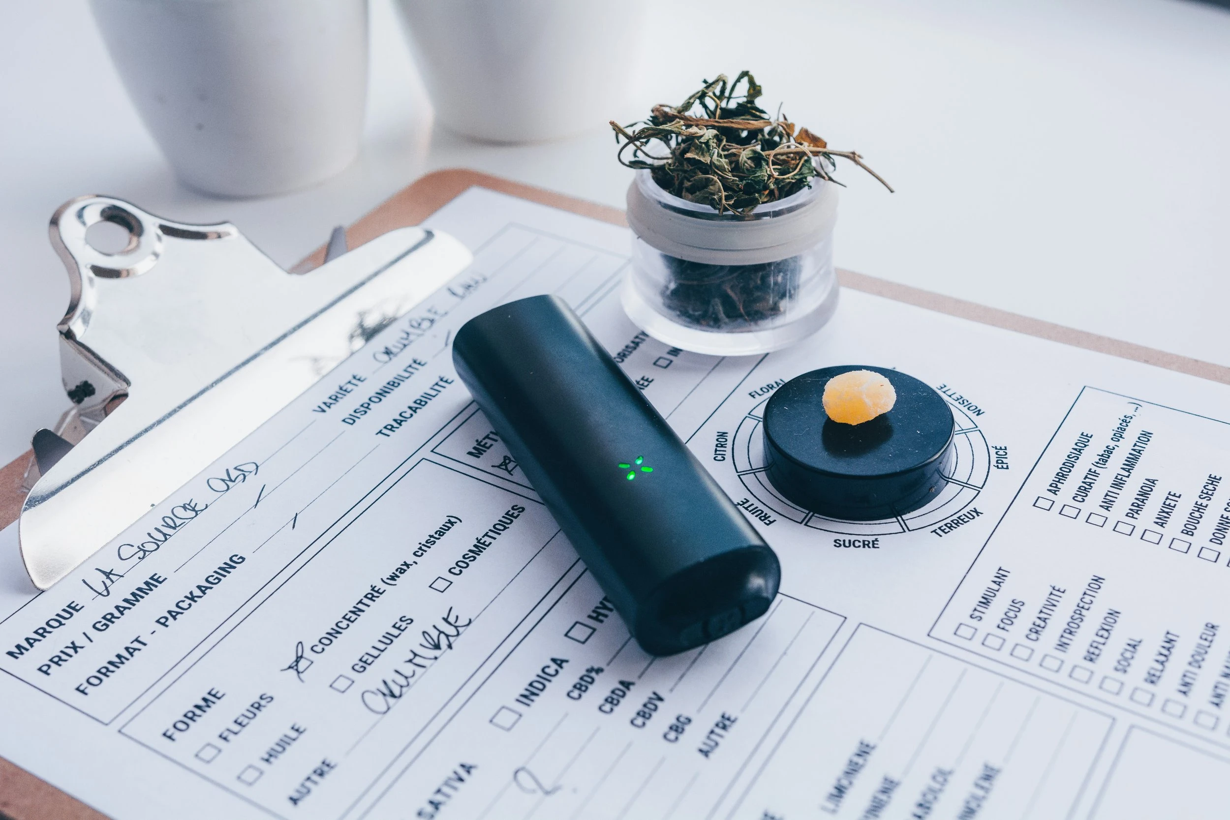 A black PAX dried herb vaporizer resting on a clipboard full of answered questionaries and an open jar of dried herb.