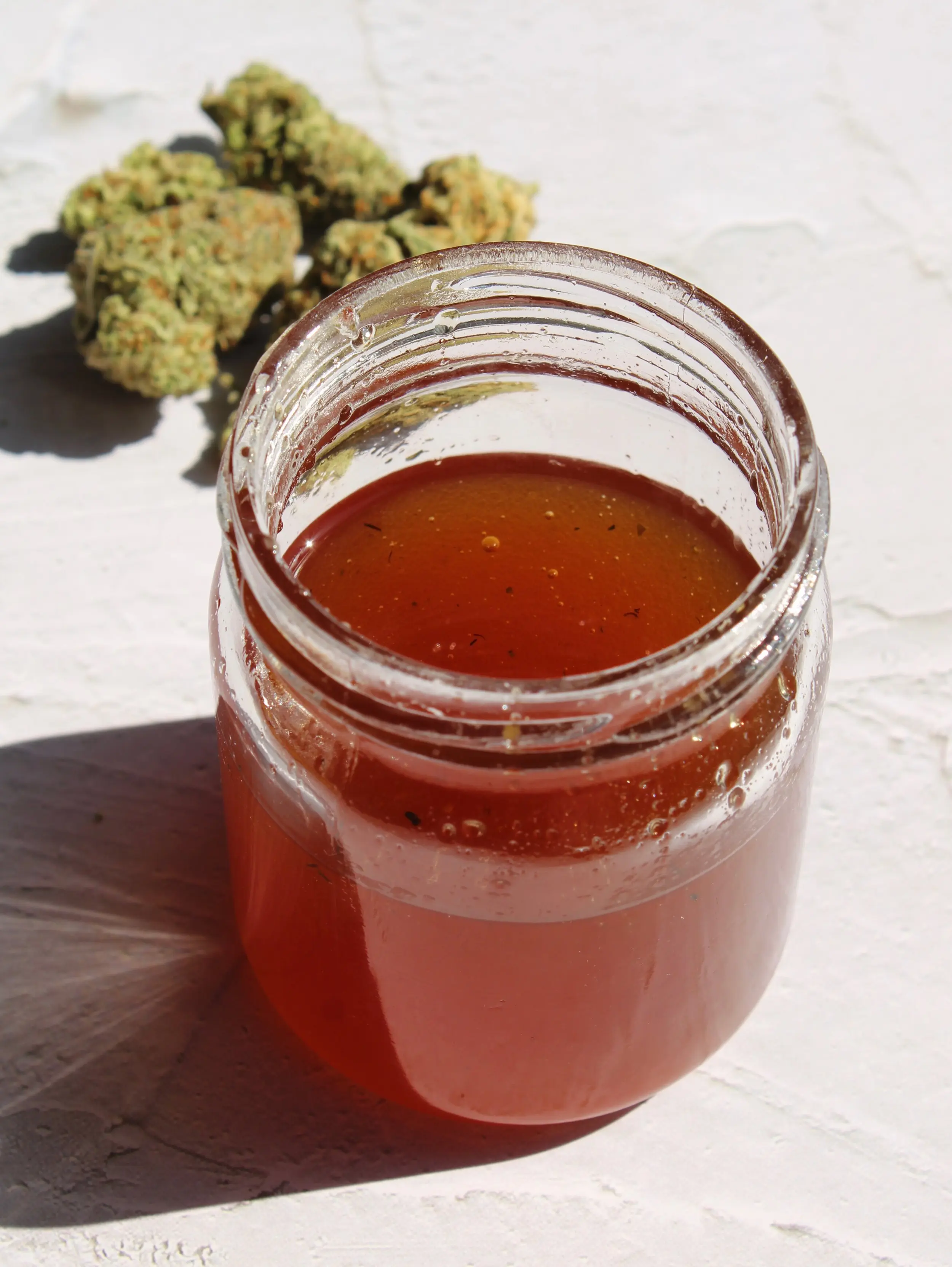 A jar of infused honey sitting on a marble counter. A large nugget of cannabis sits on the top left of the counter. 