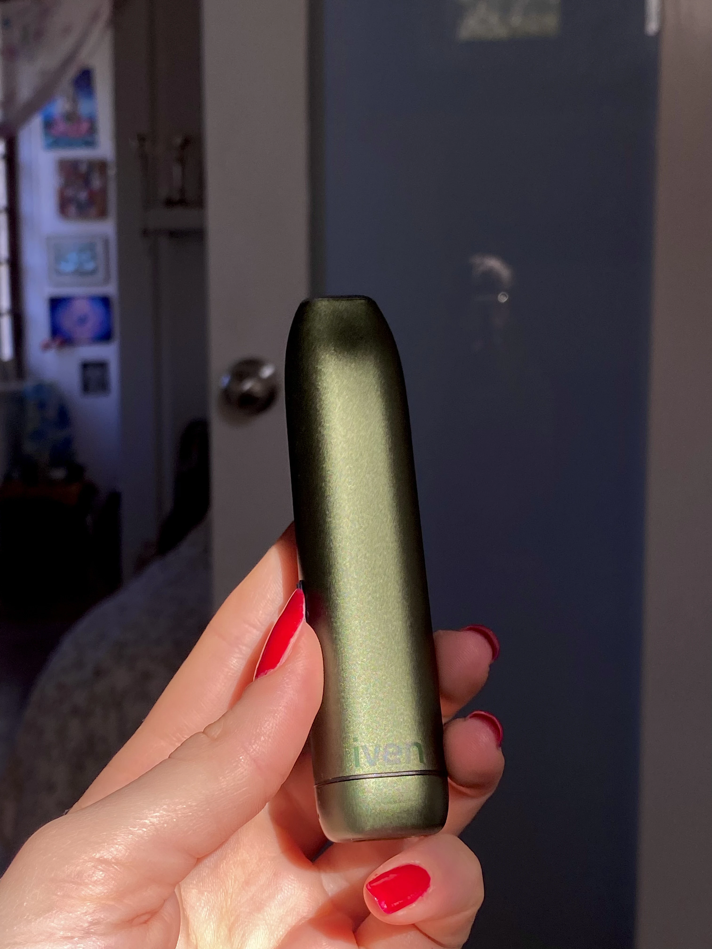 A hand with long red fingernails holding up the camp green Iven dry herb vaporizer in the sunlight with a doorway in the background.