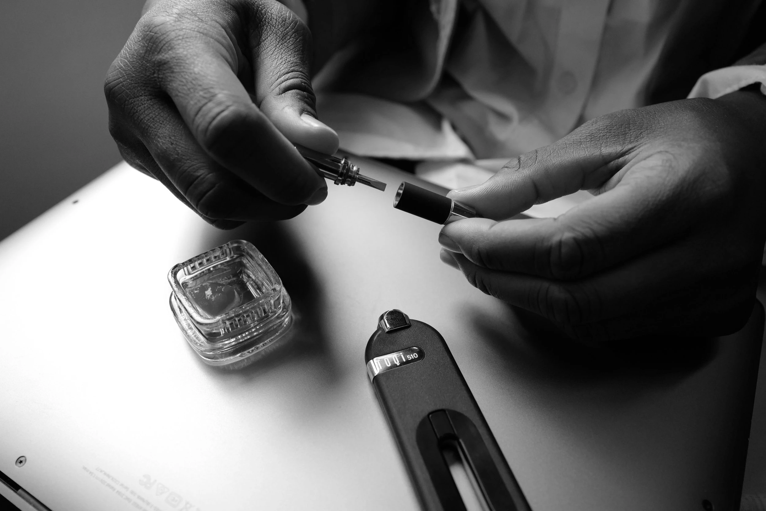 A black and white photo of a Toqi wireless vape adaptor rests on a table while a hand inserts a cartridge to the mouthpiece.