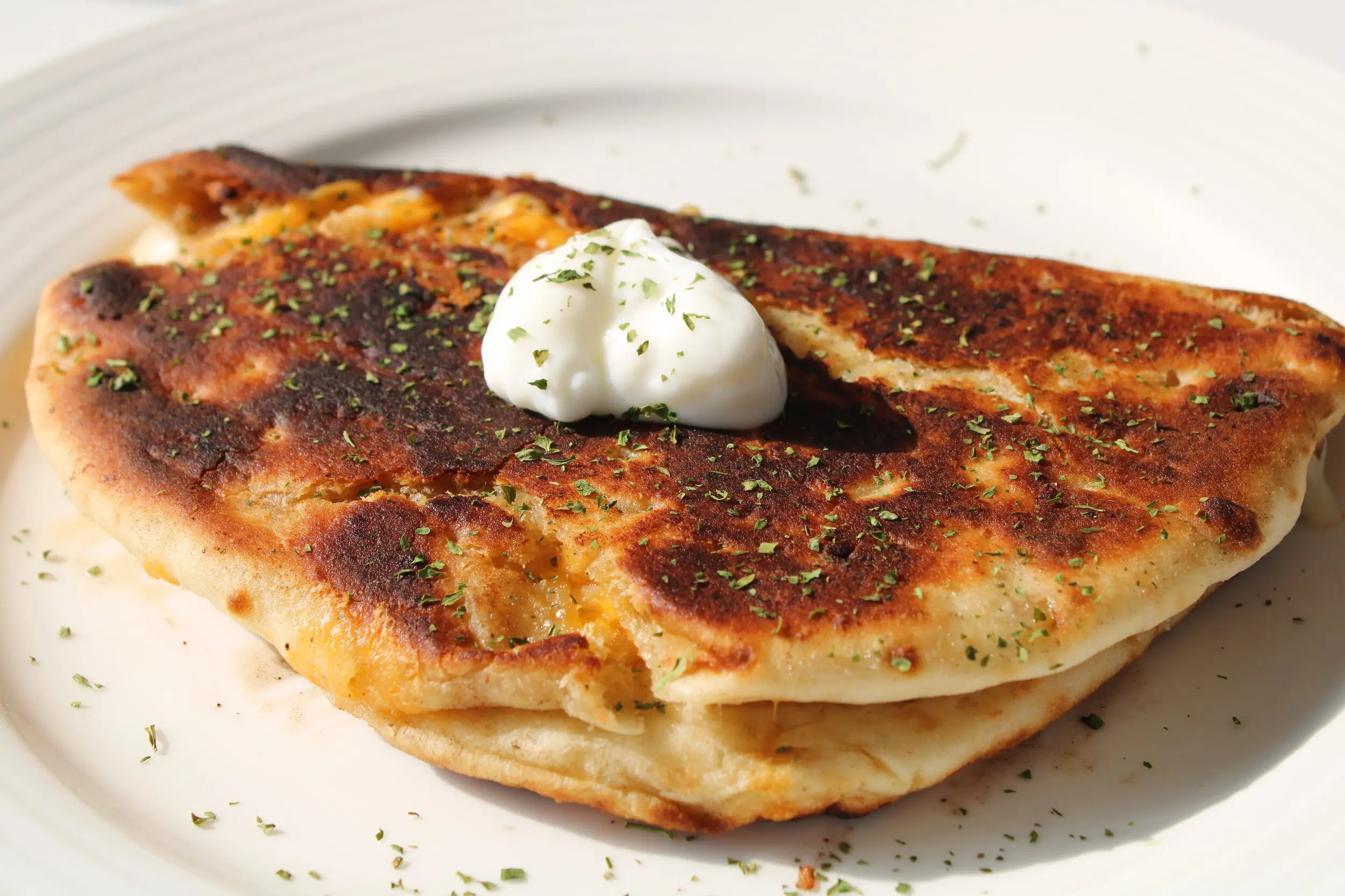 a naan grilled cheese resting on a cream coloured plate with a dollop of sour cream on top. Shaved over the the naan and the plate is oregano.