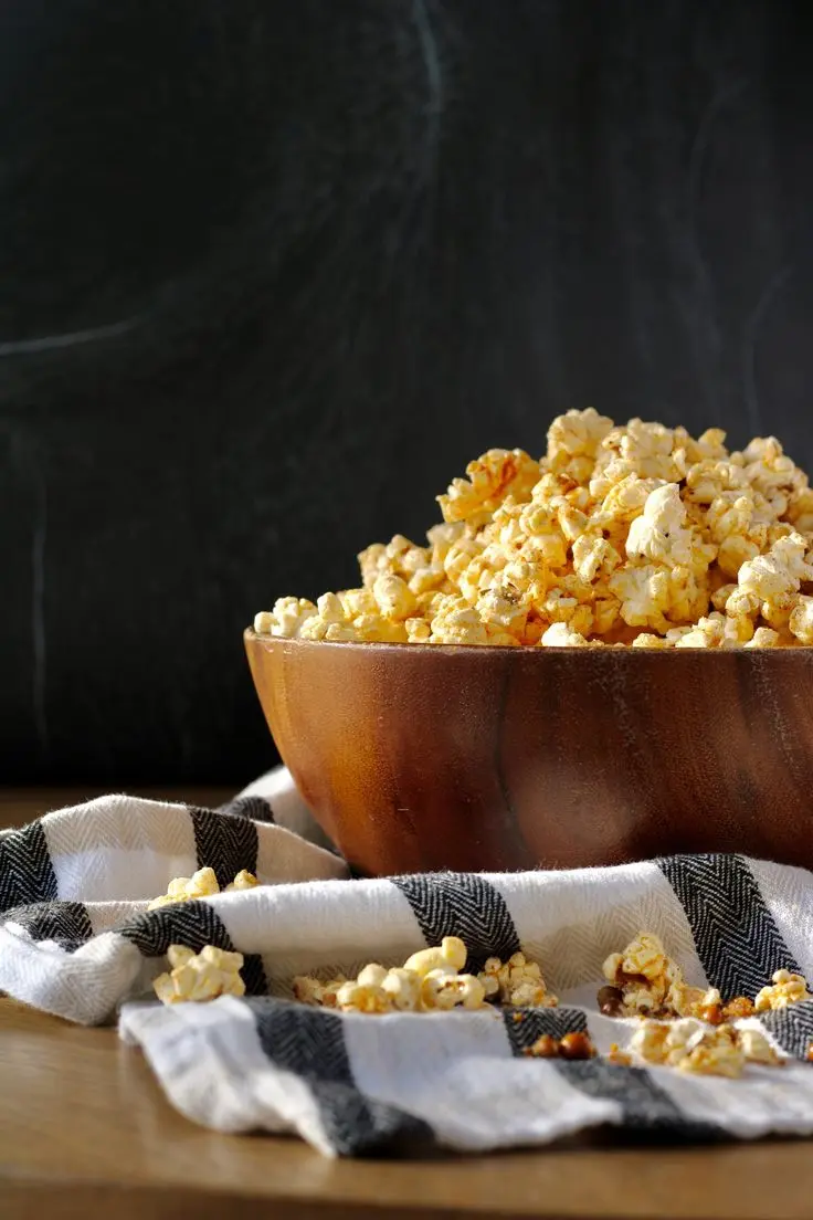 a dark wooden bowl overflowing with buttery popcorn on top of a white and black striped tea towel which has scattered pieces of popcorn on it.