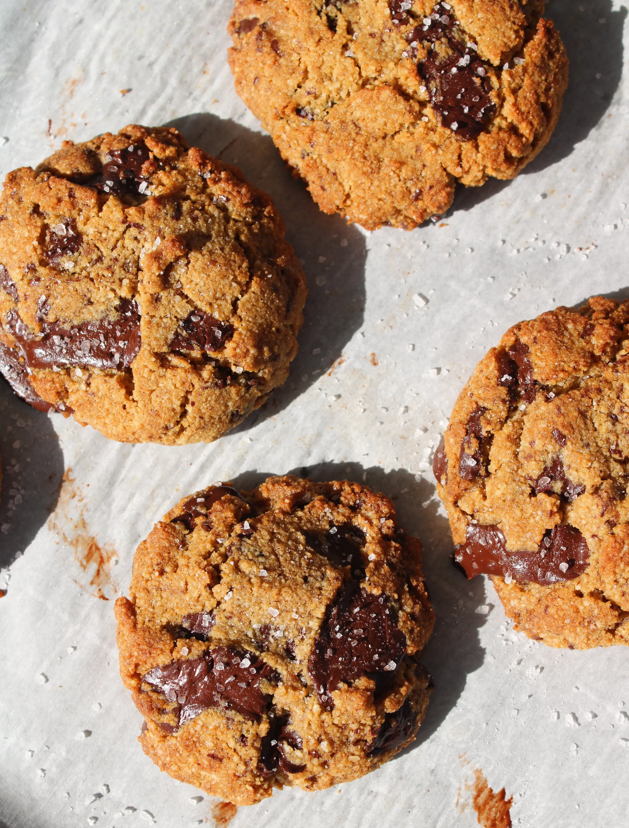 4 round chocolate chunk cookies with a sprinkle of large flaked sea salt on them. They sit on a marble counter.