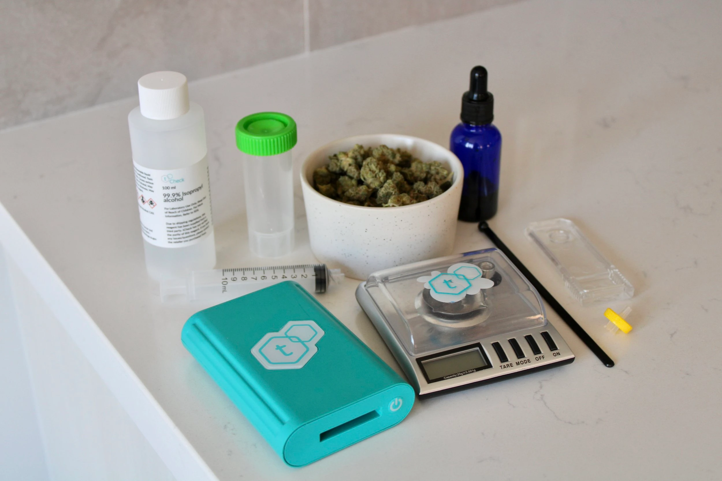 A turquoise blue t-check potency tester resting on a marble countertop next to a tcheck branded scale and its accompanying tools, cannabis nuggets and dropper bottles.