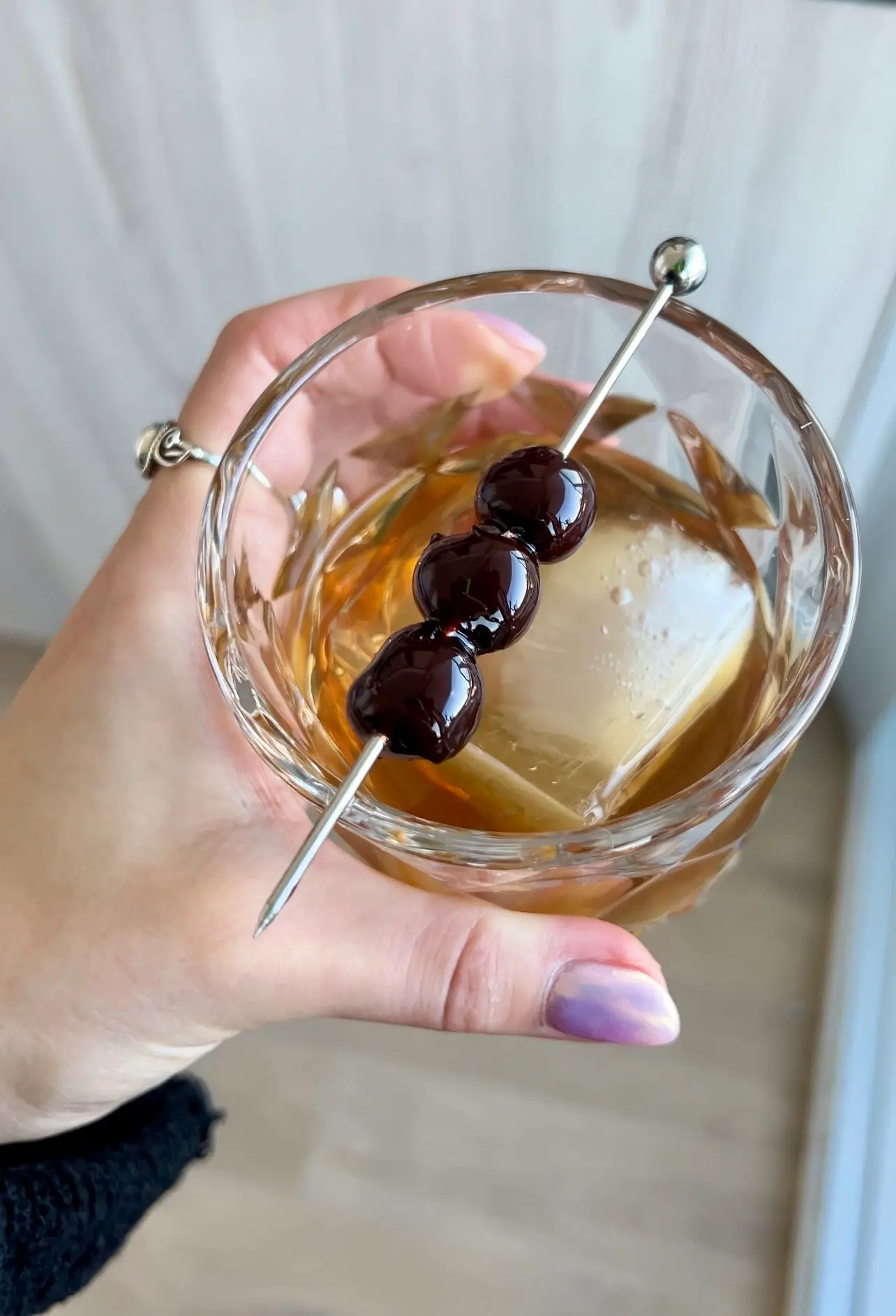 a hand holding a rocks glass full of an amber liquid. One square ice cube rests in the centre while a toothpick with 3 black berries sits on the top of the glass as a garnish.