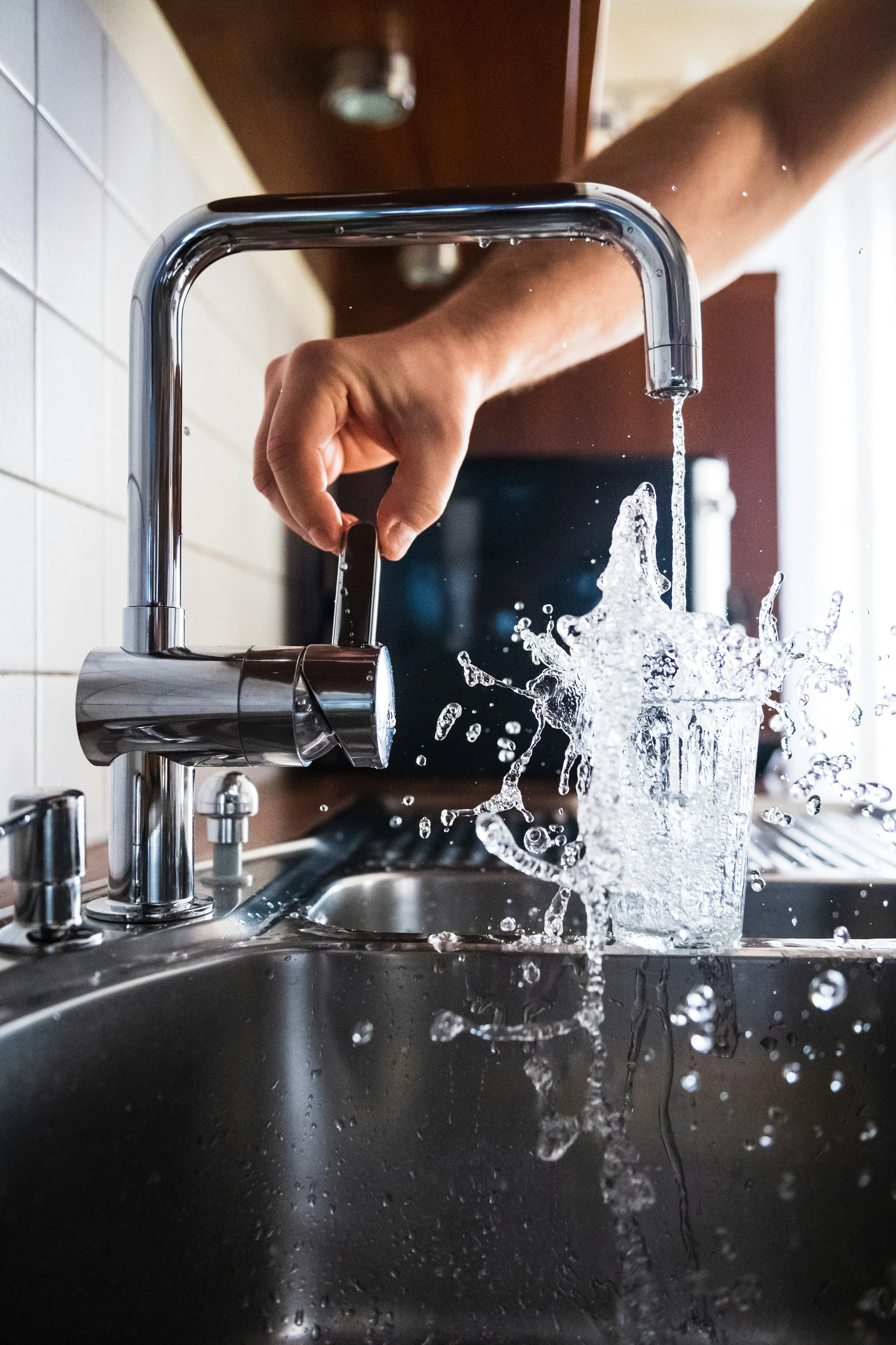 A hand turning on a stainless steel sink which fills up a glass jar completely and splashes water all over the counter. 