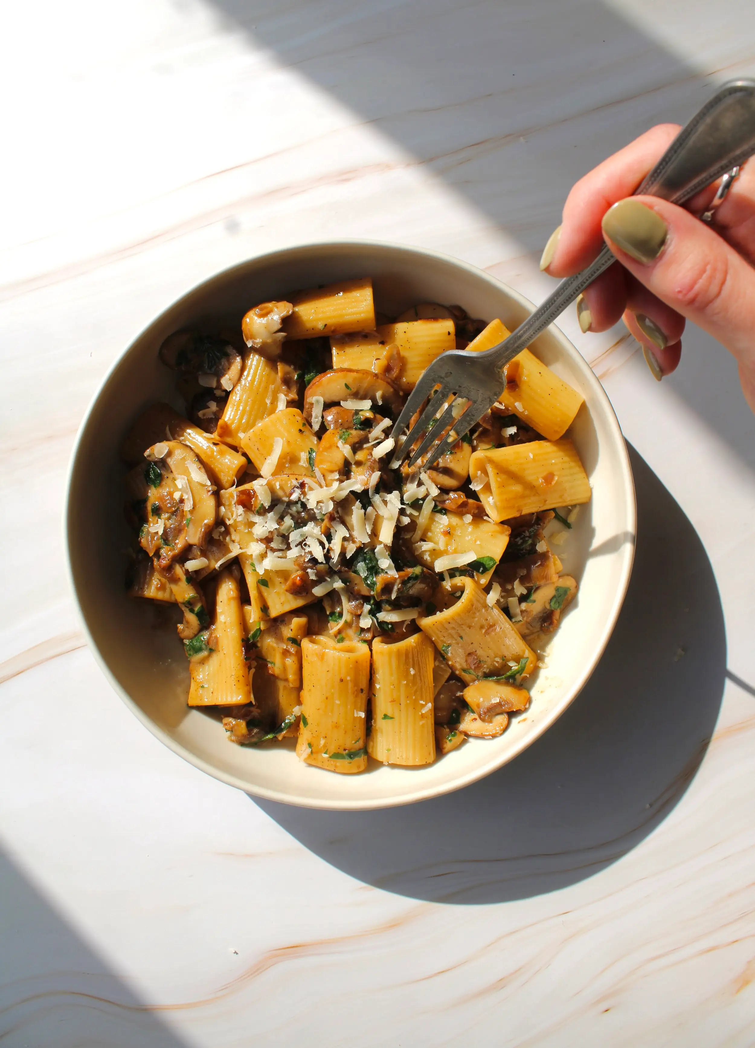 an aerial shot of a large bowl of rigatoni pasta that is topped with herbs and parmesan cheese. On the right side of the photo is a hand with a fork that is reaching for the pasta.