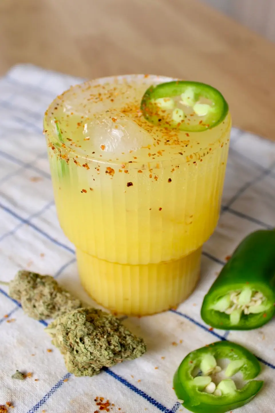 A clear, ridged cocktail glass with the mango margarita and a few ice cubes in it filled to the brim. On the striped cloth under the margarita glass are sliced jalapeños and a nug of cannabis flower.