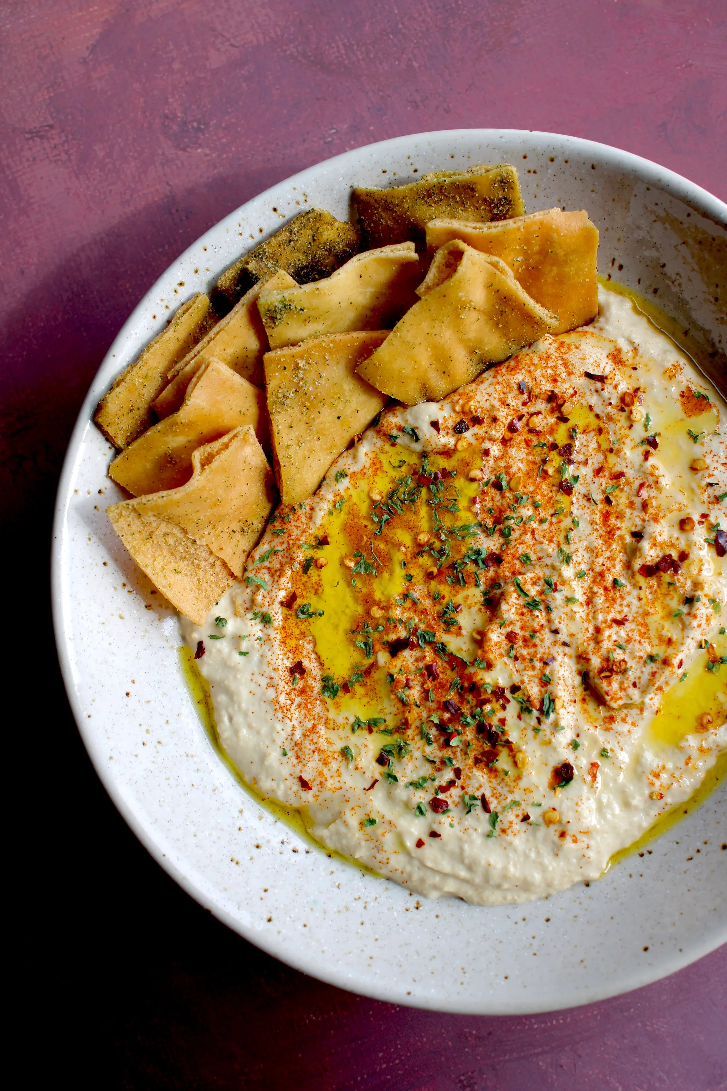 an aerial shot of a beige plate of hummus. On the left side is a portion of tortilla chips and the dish is garnished with red and green herbs and spices.