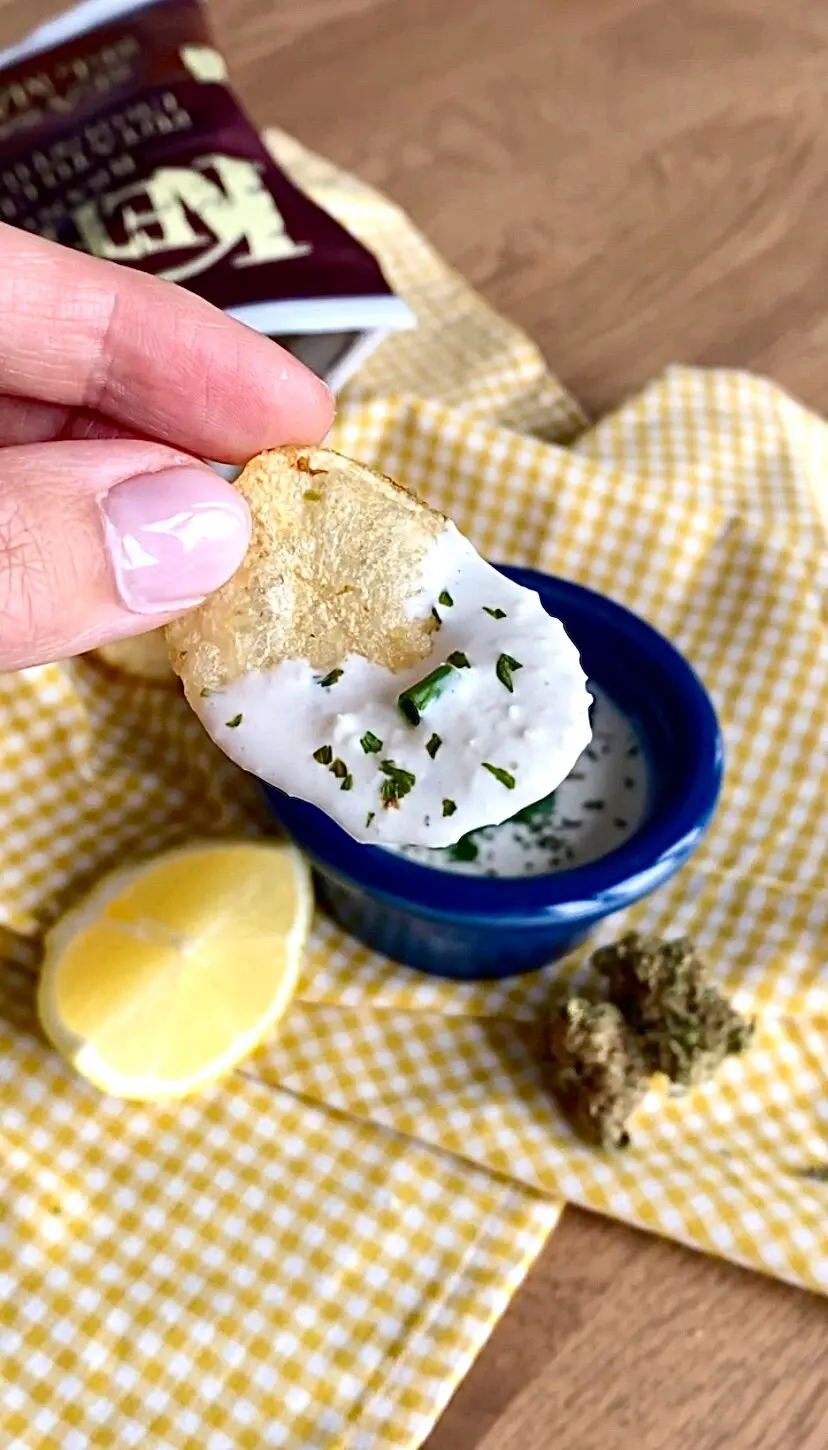 A chip that has been clearly dipped in the blue bowl of onion dip below it, with a lemon wedge and cannabis flower nugget right next to it. 