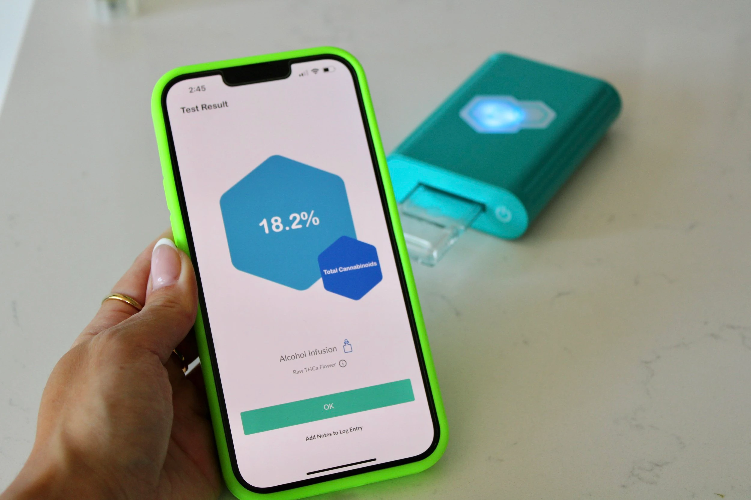 An iPhone with a green case displaying an 18% result from using the turquoise cannabis tCheck machine behind it. 