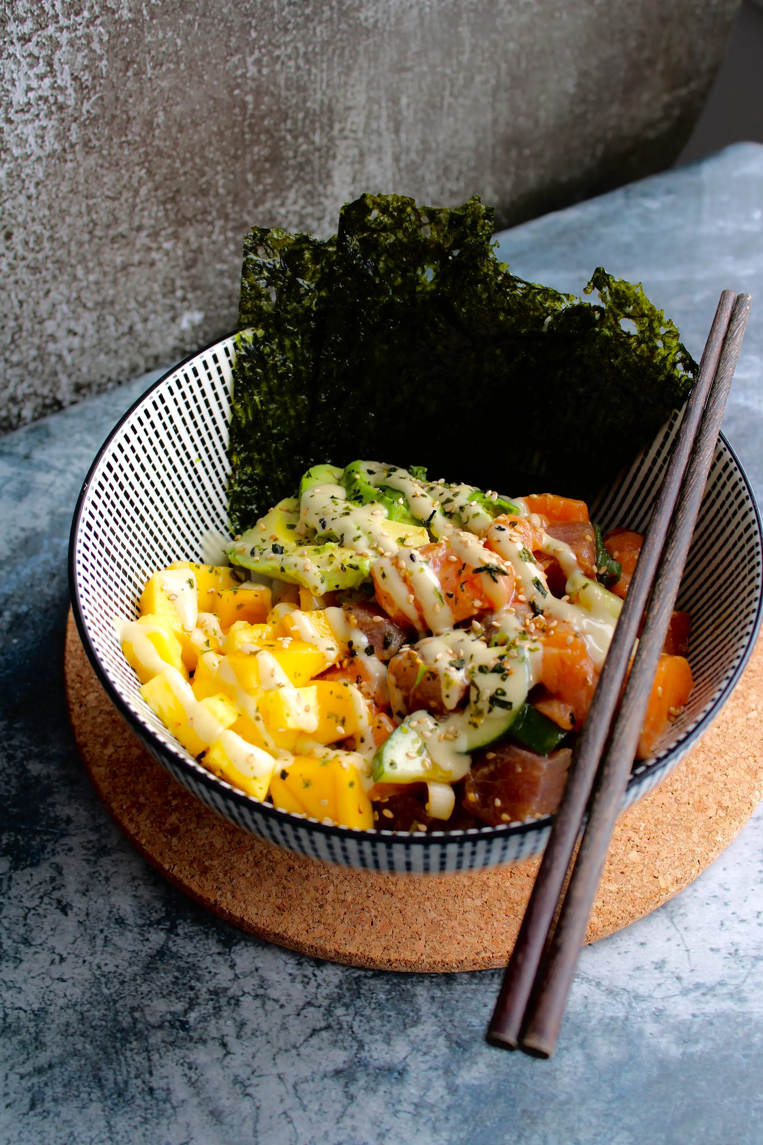 A striped bowl full of poke fixings like avocado, mango, seaweed and salmon. A pair of chopsticks rests on the side of the bowl.