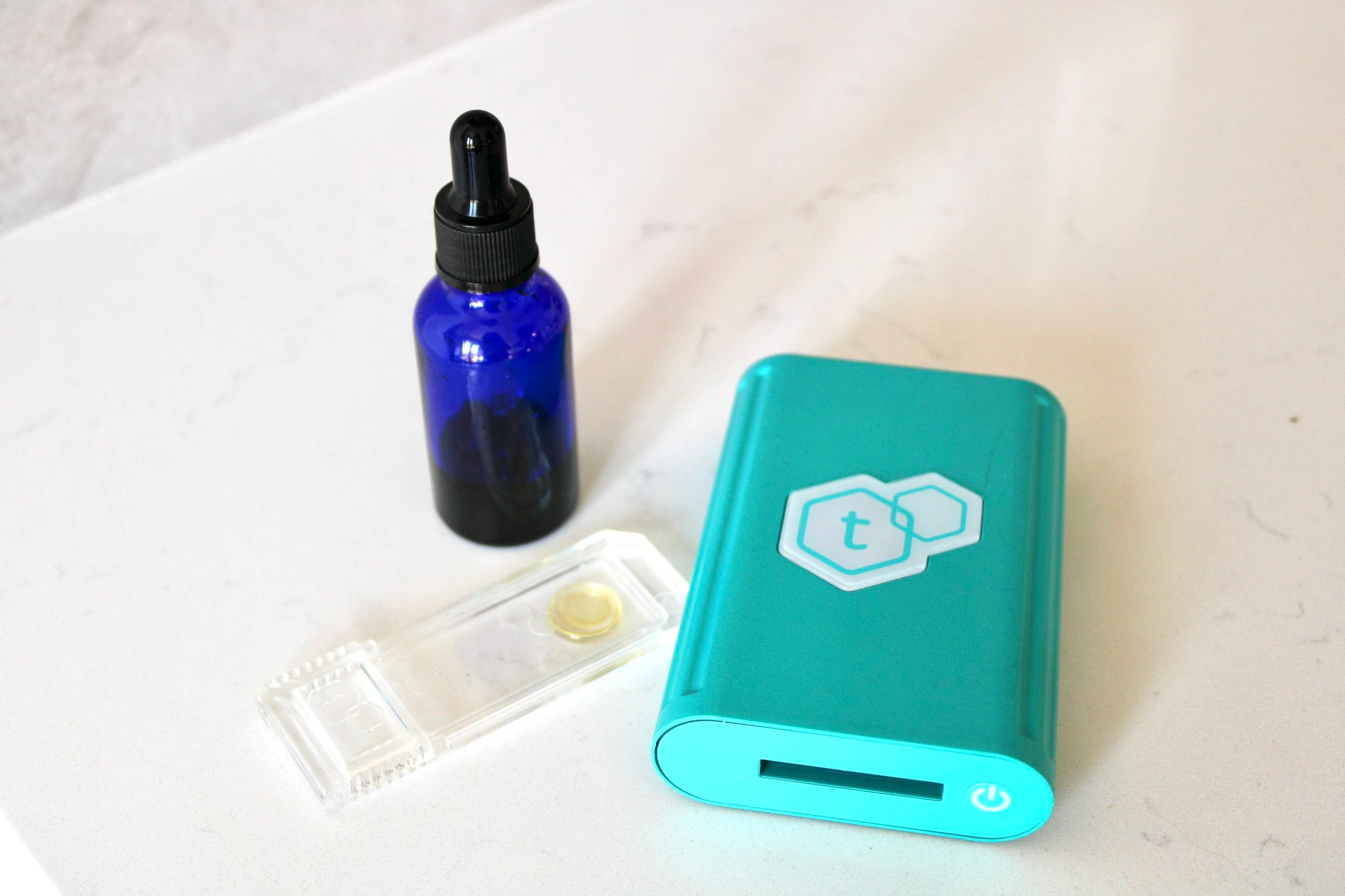 A turquoise tcheck potency device resting on a marble counter next to a blue dropper bottle with a black lid, as well as a clear testing strip with a dab of oil resting in the centre of it.