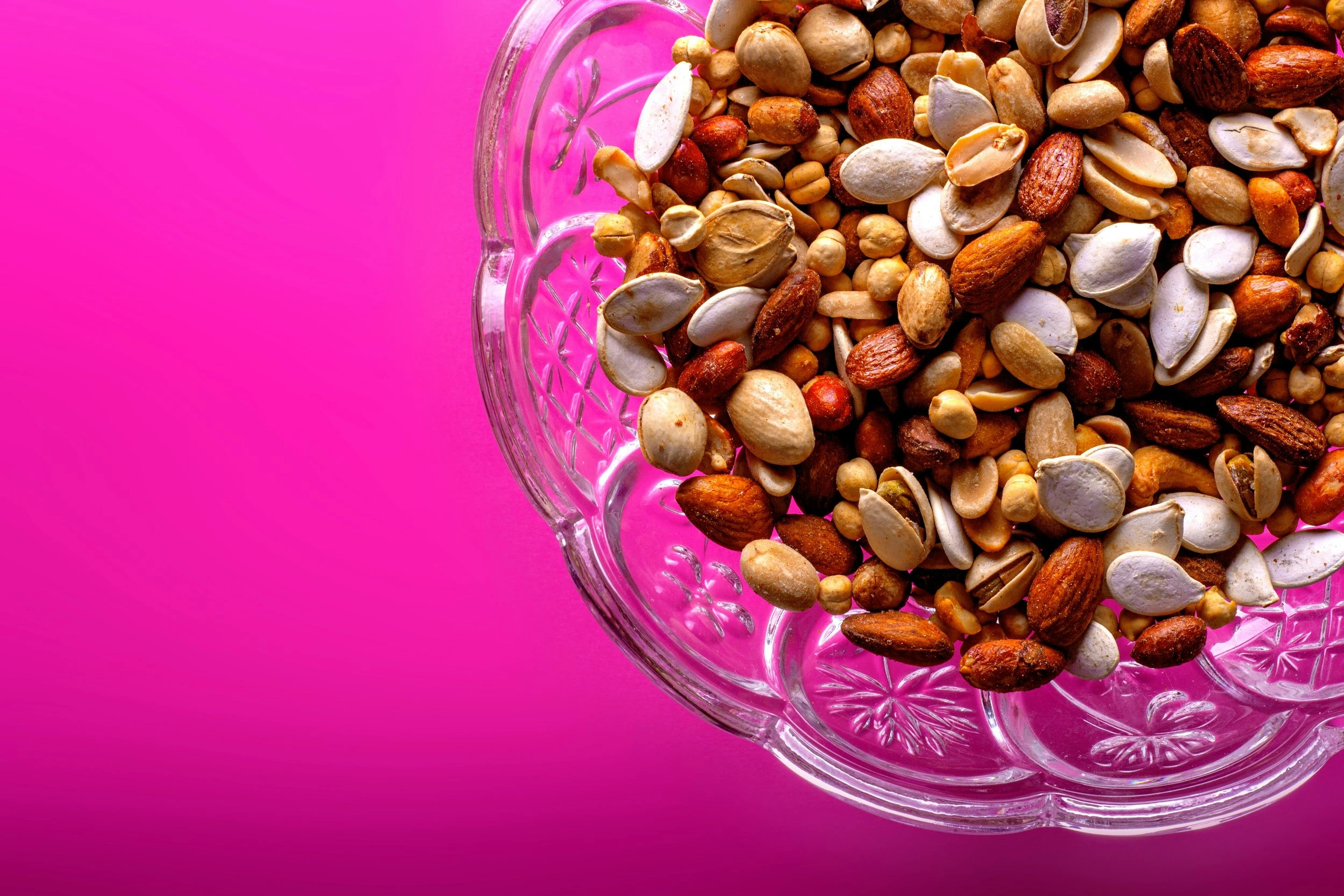 A glass bowl of trail mix resting against a vibrant pink background. 