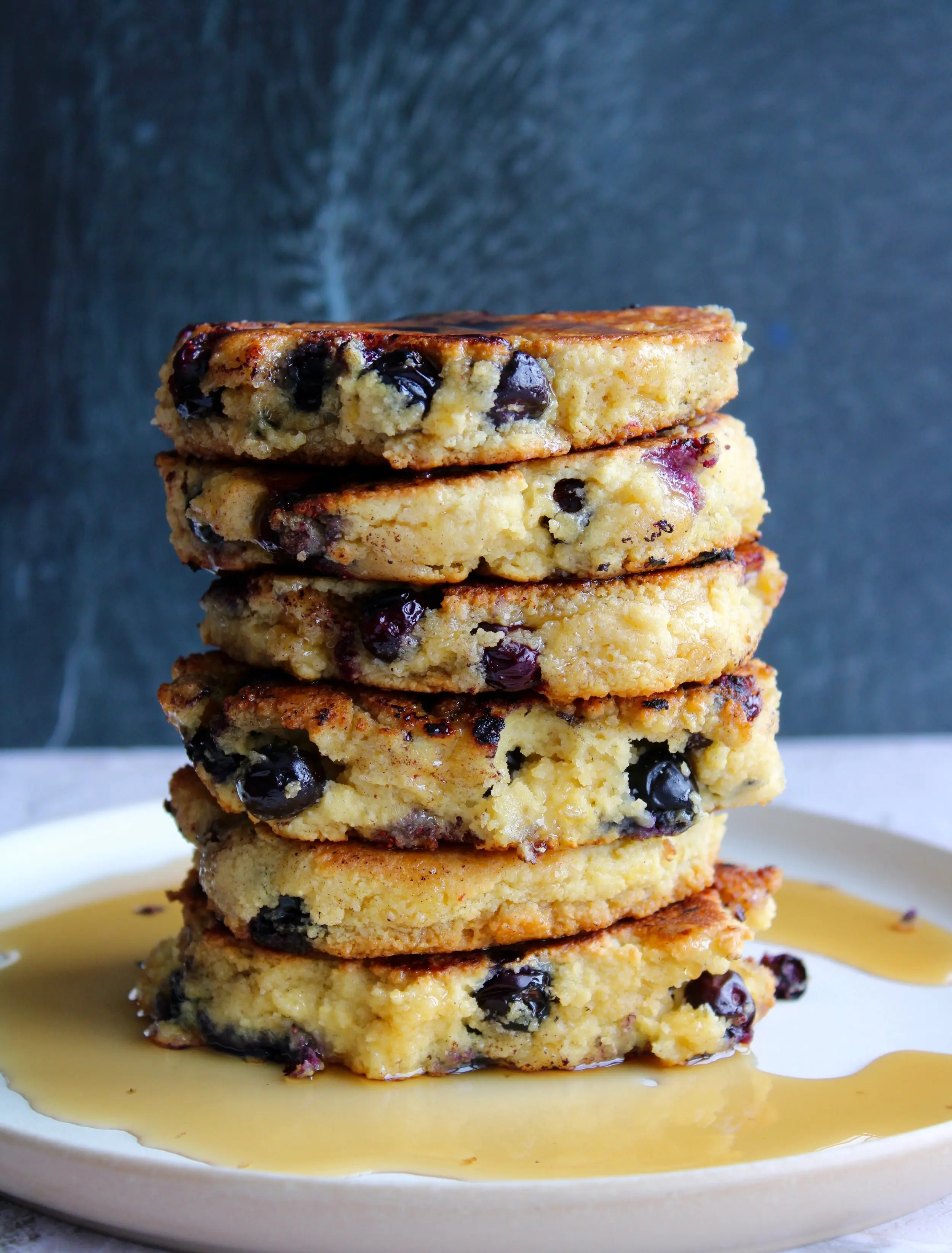 a six stack pancake stack on top of a cream plate. The pancakes are drizzled with maple syrup which catches at the base of the plate.