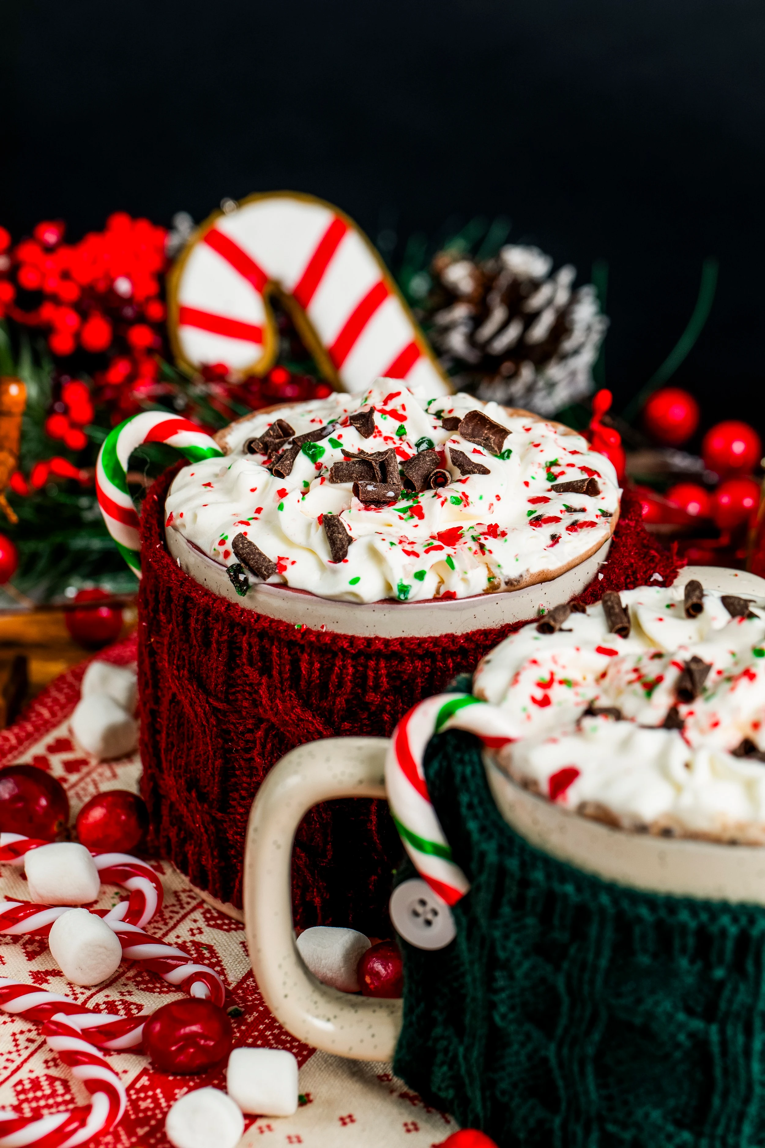 Two mugs in red and green knitted cozies full to the brim with cannabis infused hot chocolate and topped with whipped cream, chocolate shavings and red and green sprinkles.