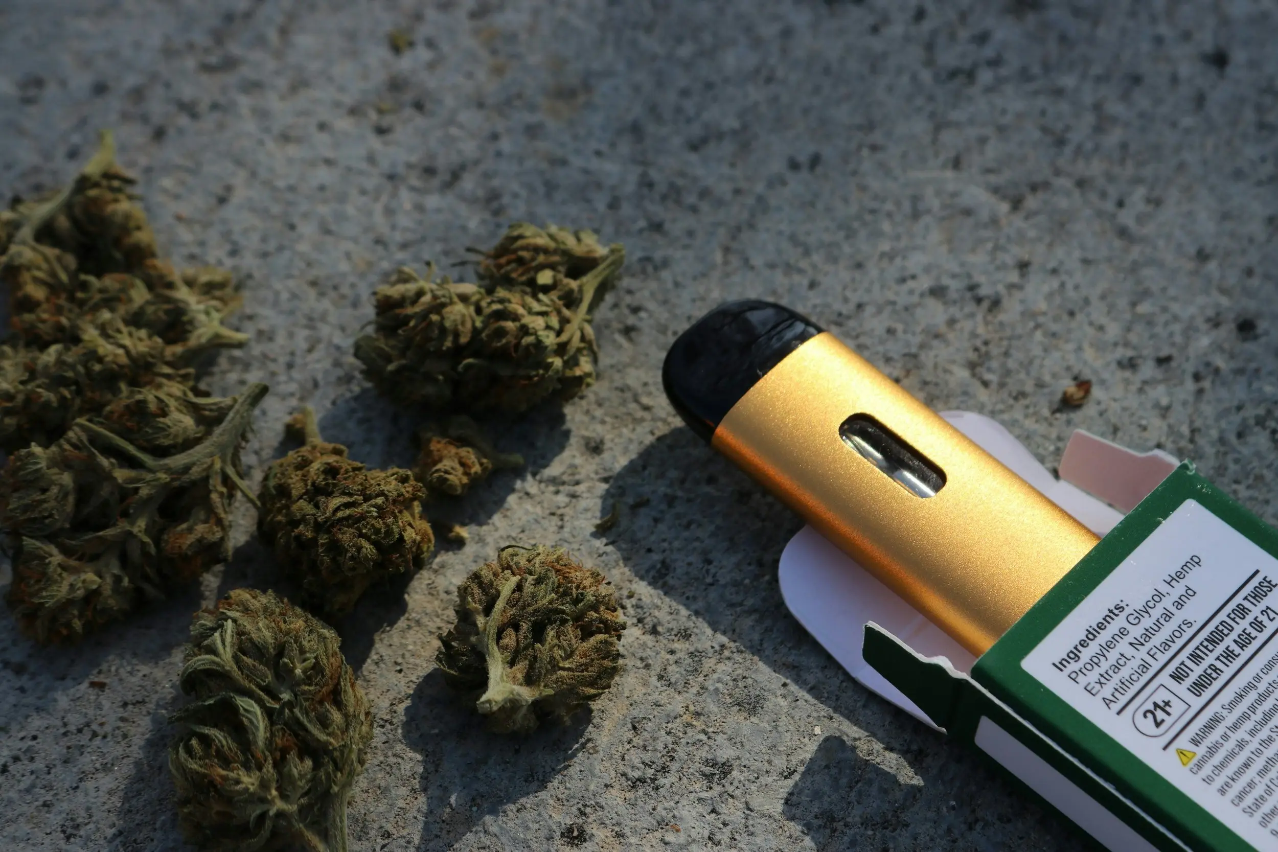 A gold vape pen rests half out of its green packaging, next to a handful of cannabis nuggets. 