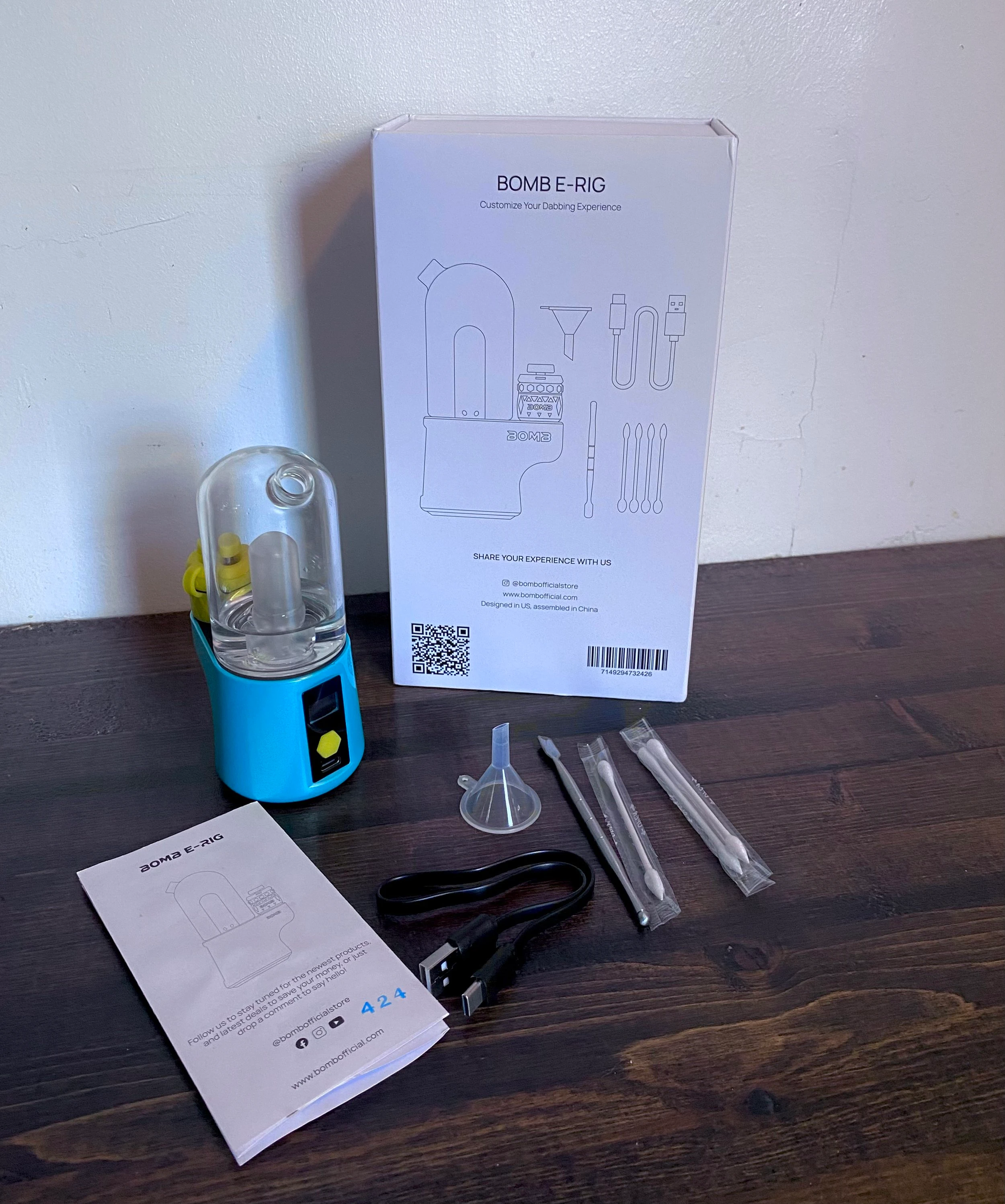 The Bomb Electronic dab rig resting on a dark wooden countertop with all of the accompanying tools in the packaging including Q-tips, a strainer, the charging chord and p