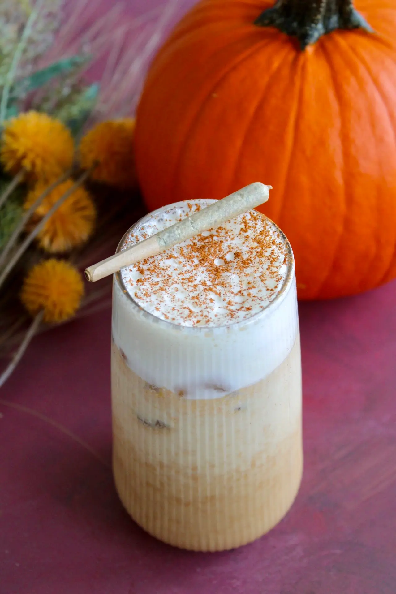 a clear glass filled to the top with iced pumpkin spiced latte topped with cold foam, garnished with a joint on the rim. Behind it lies a pumpkin and some fall coloured flowers.