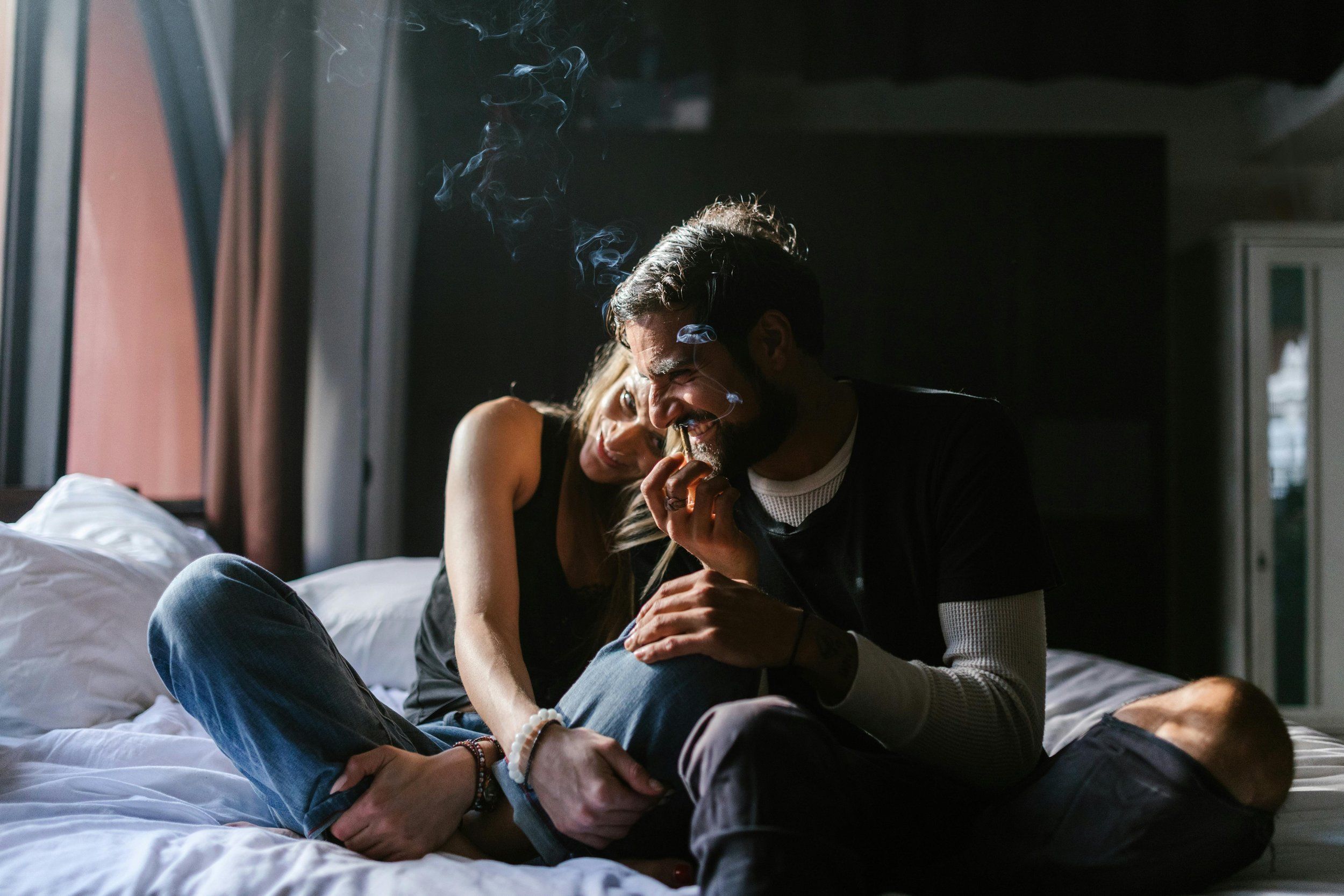 A young couple sitting cross legged on a bed, smiling and sharing a cannabis joint. 