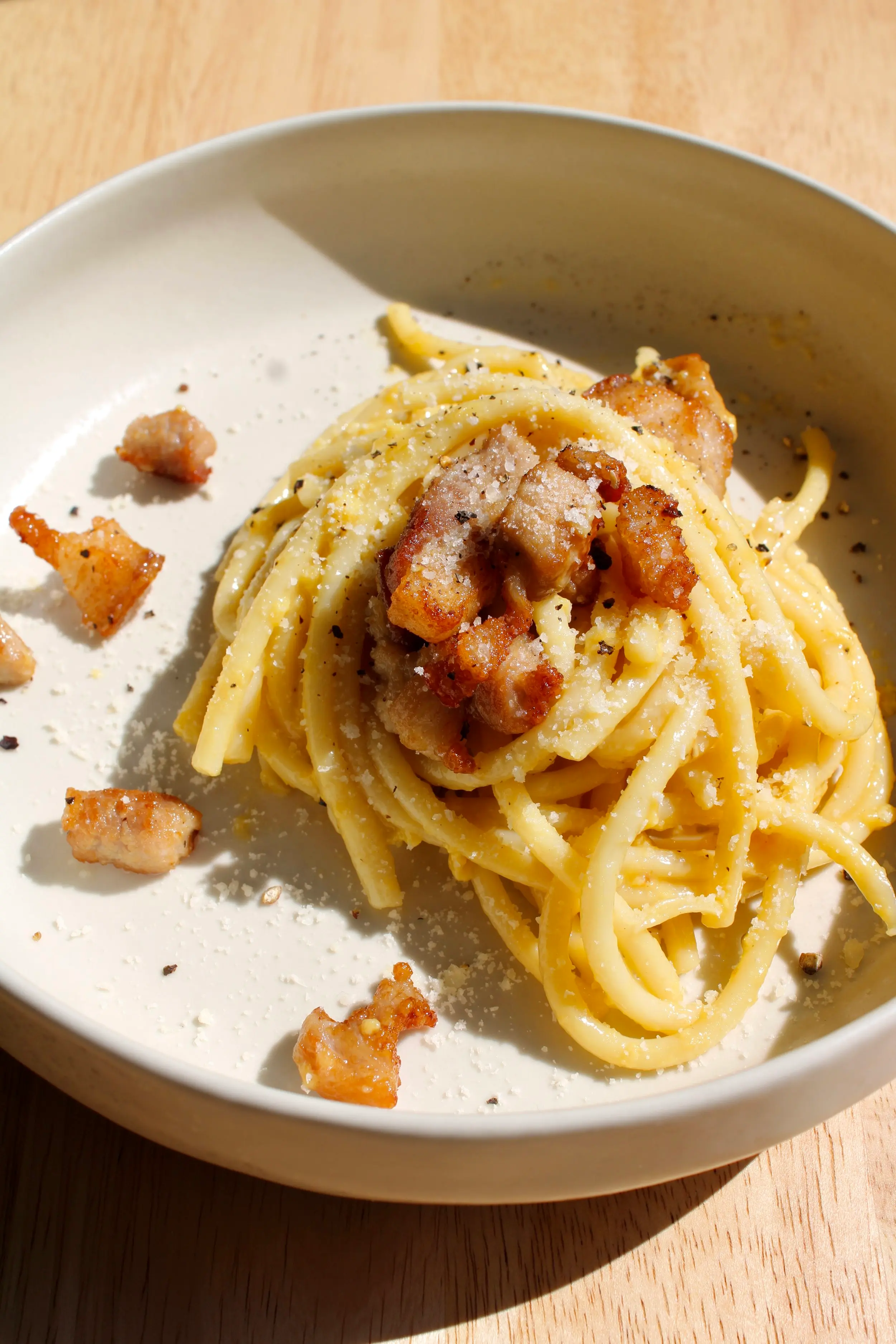 a cream coloured plate of carbonara pasta with bits of pork belly and parmesan cheese on top.