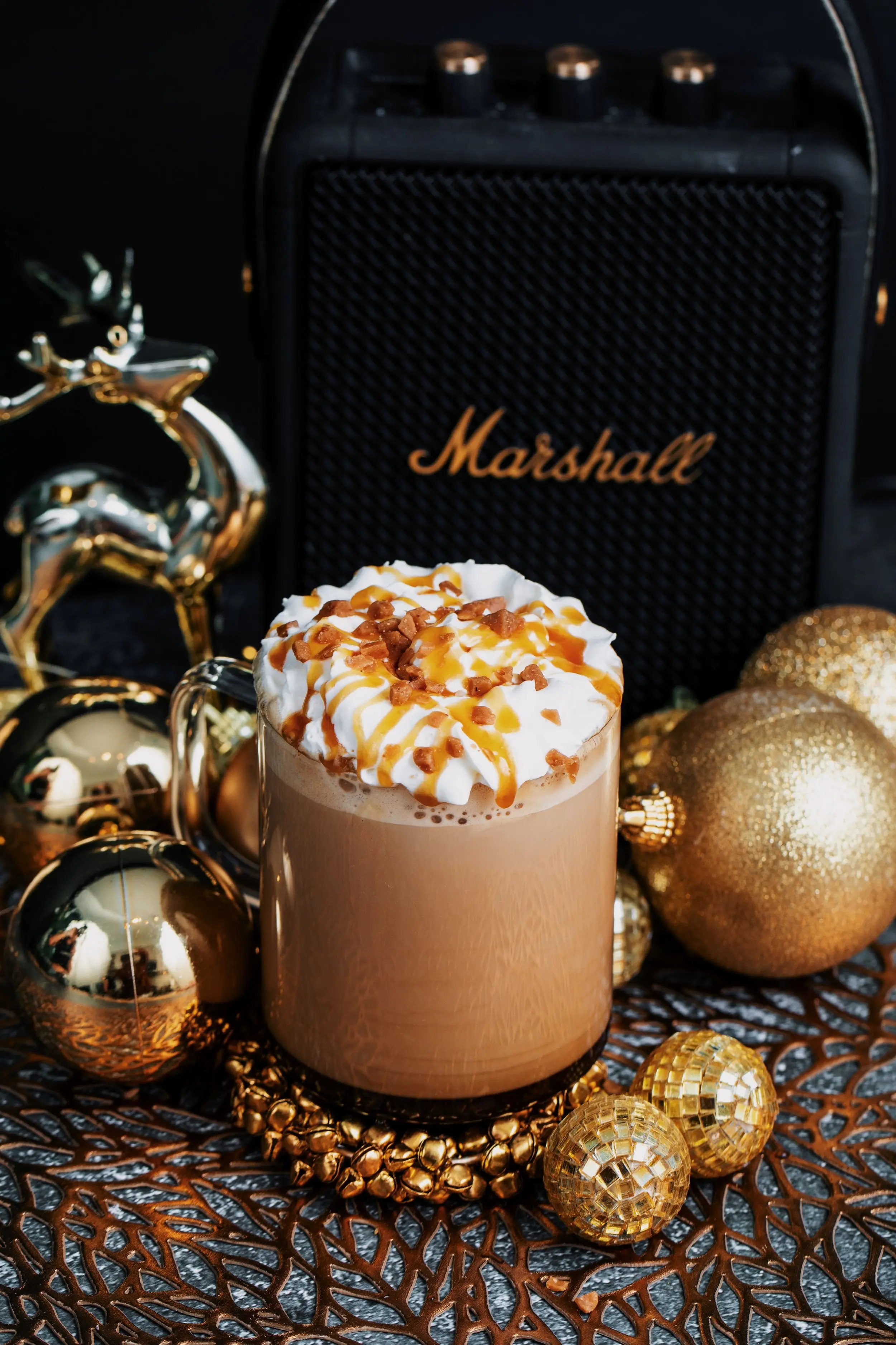 A glass mug containing a latte, topped with whipped cream and a caramel drizzle. It sits on a bed of gold Christmas decorations in front of a Marshall speaker. 