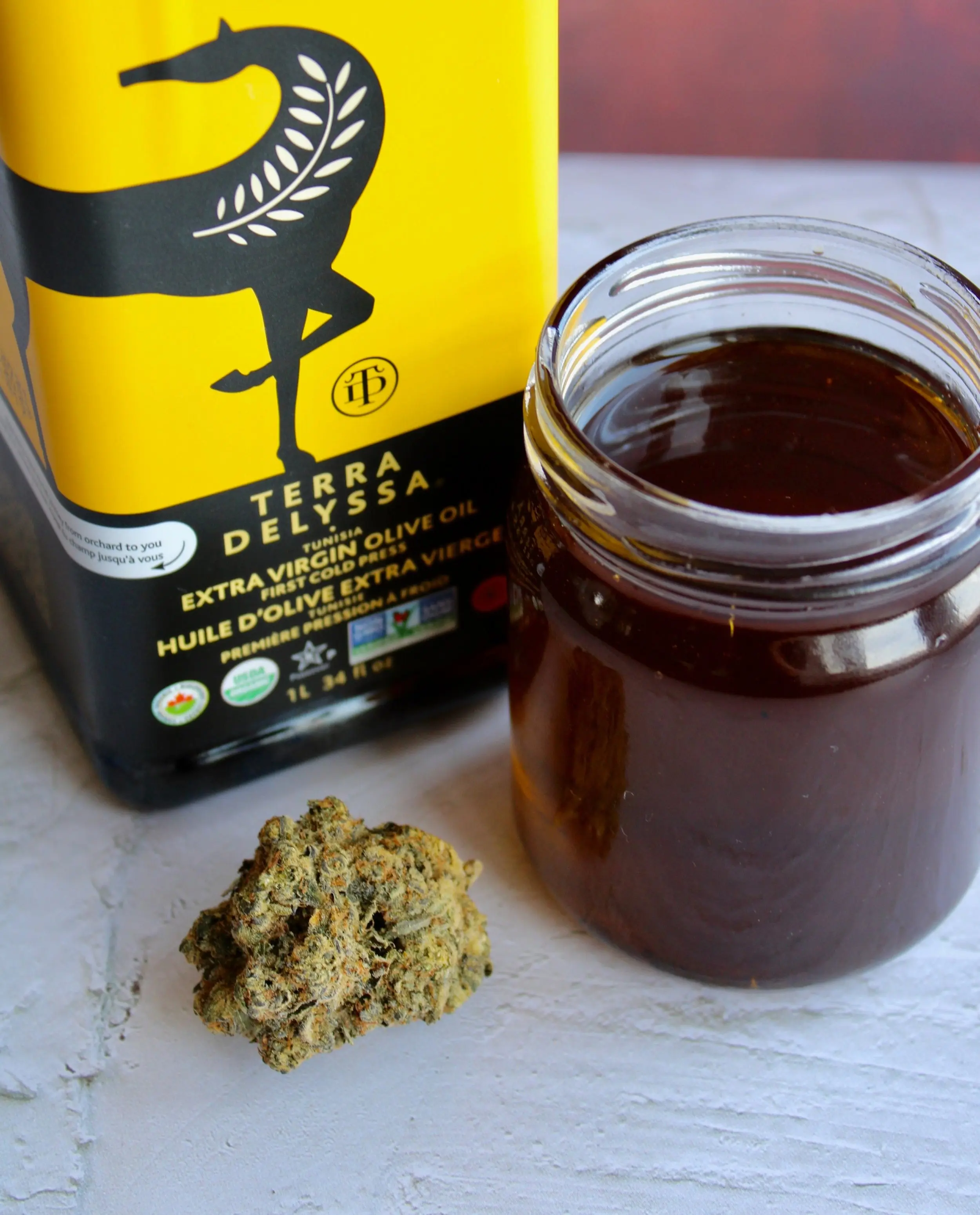 A clear glass jar filled almost to the top with dark cannabis oil. On the front left side of the jar is a large nugget of cannabis, and on the back left side is a large bottle of olive oil.