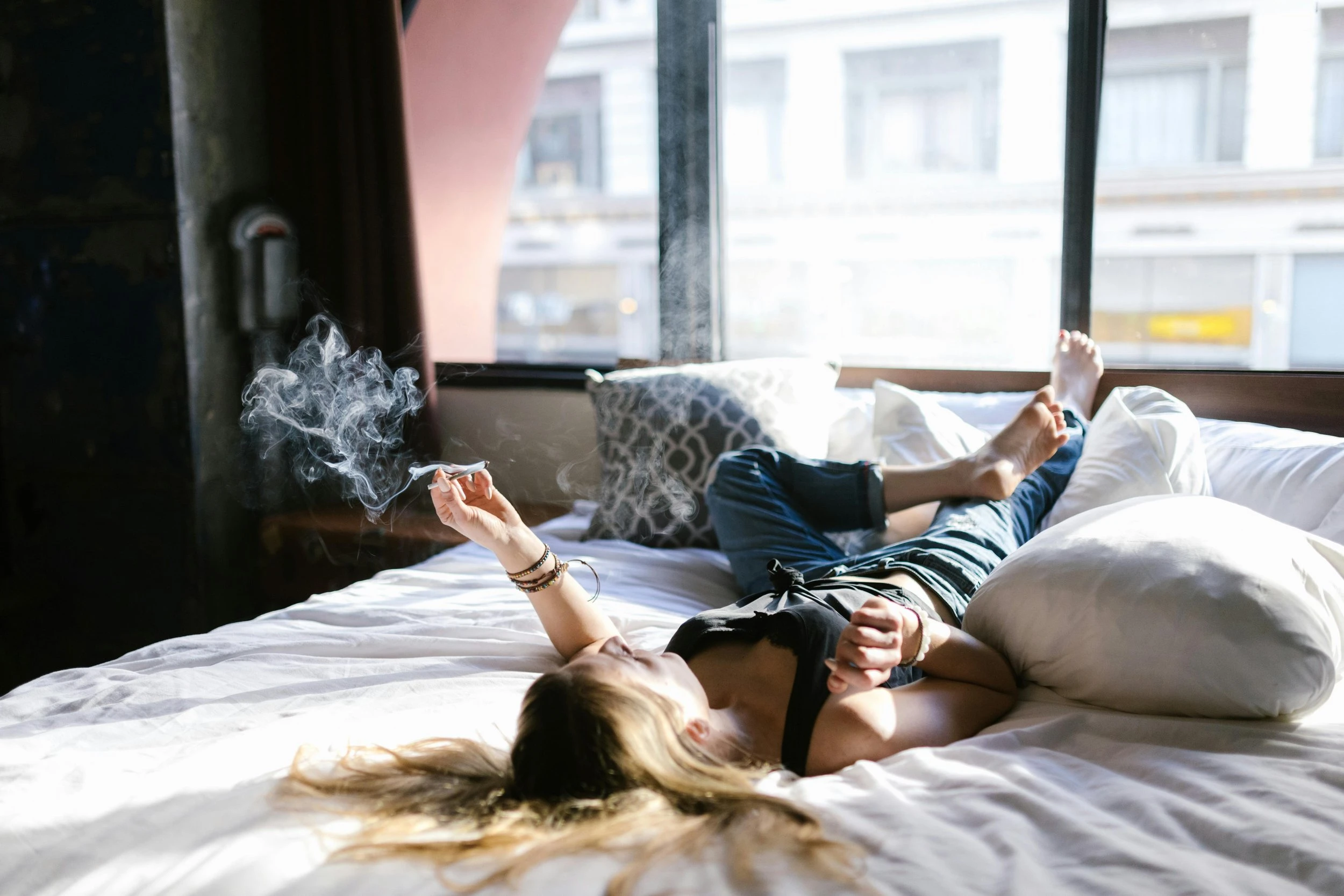 A woman lying backwards in bed, on her back, holding a lit cannabis joint. 